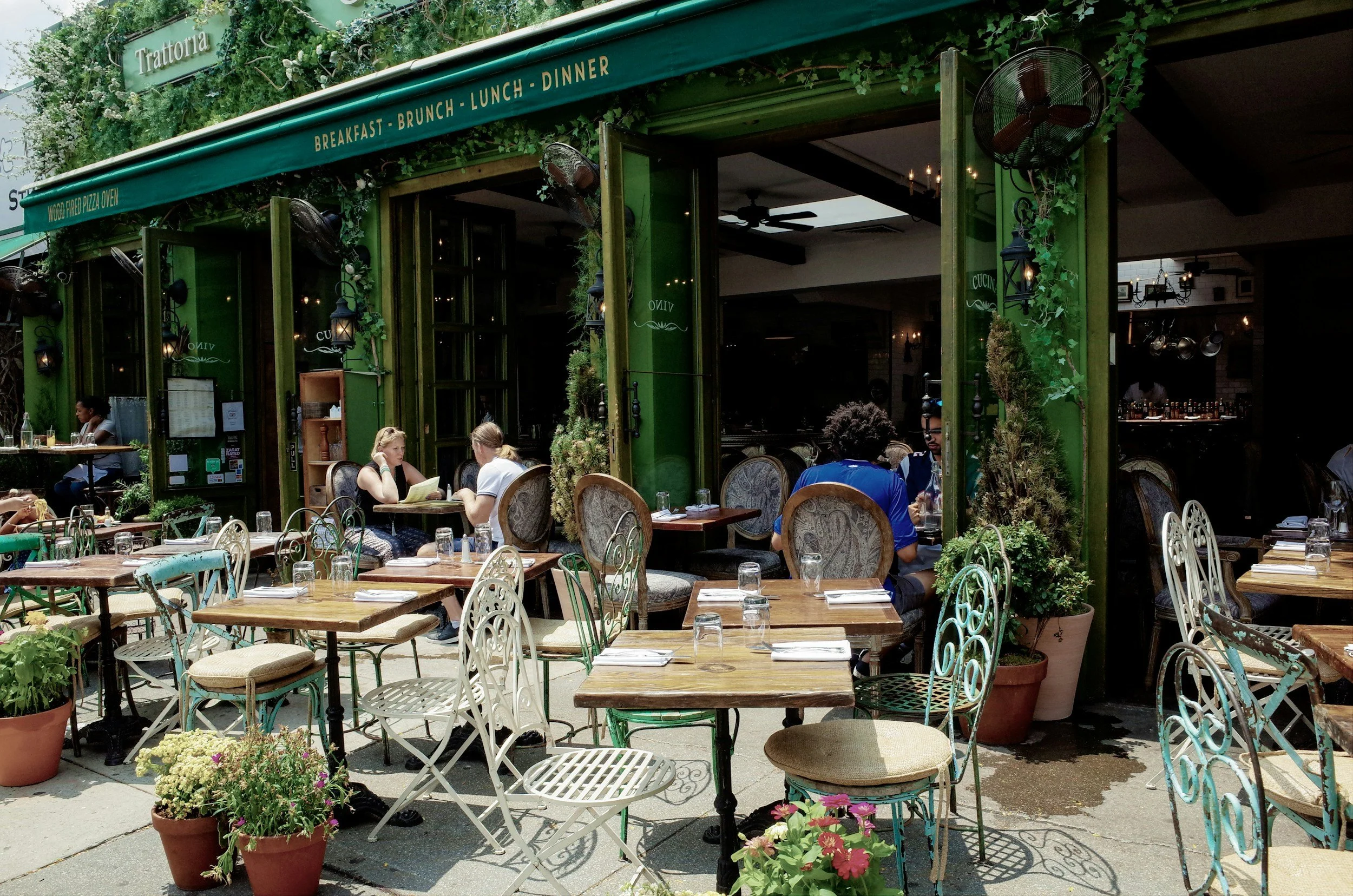 An outdoor patio of a restaurant with wooden tables and ornate metal chairs, some with cushions. Potted plants and flowers decorate the area. The restaurant has green walls and large open windows. There are people sitting at tables, some reading menus, and a bar visible inside. A green awning displays the words 'Breakfast - Brunch - Lunch - Dinner'.