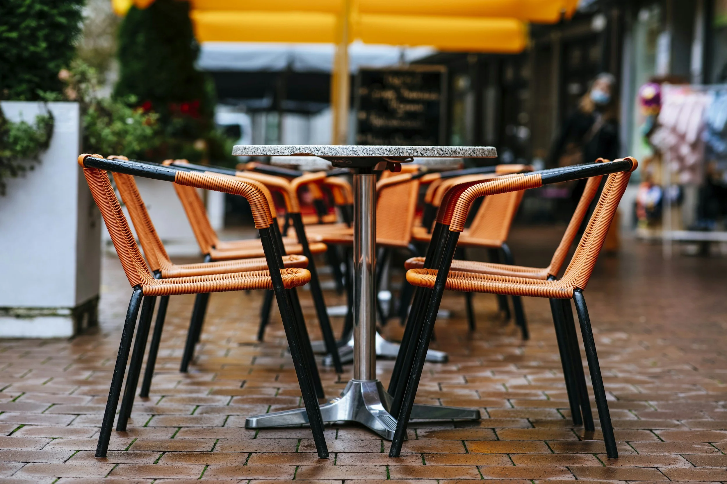 Empty outdoor cafe table with orange chairs and a gray stone tabletop on brick pavement, with a blurred background including a person wearing a mask.