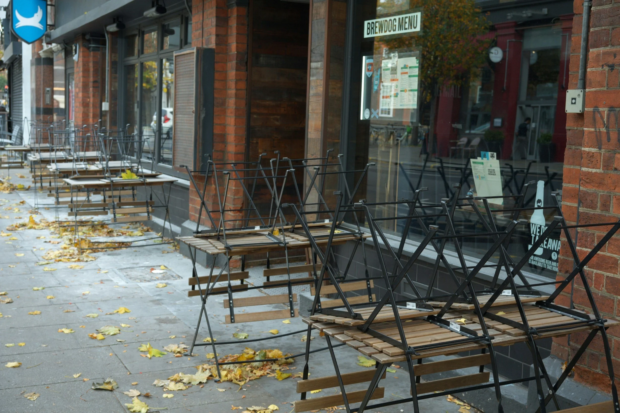 Outdoor sidewalk scene with empty tables and chairs outside a restaurant, fallen autumn leaves on the ground, brick building, and a window displaying a menu and signs.