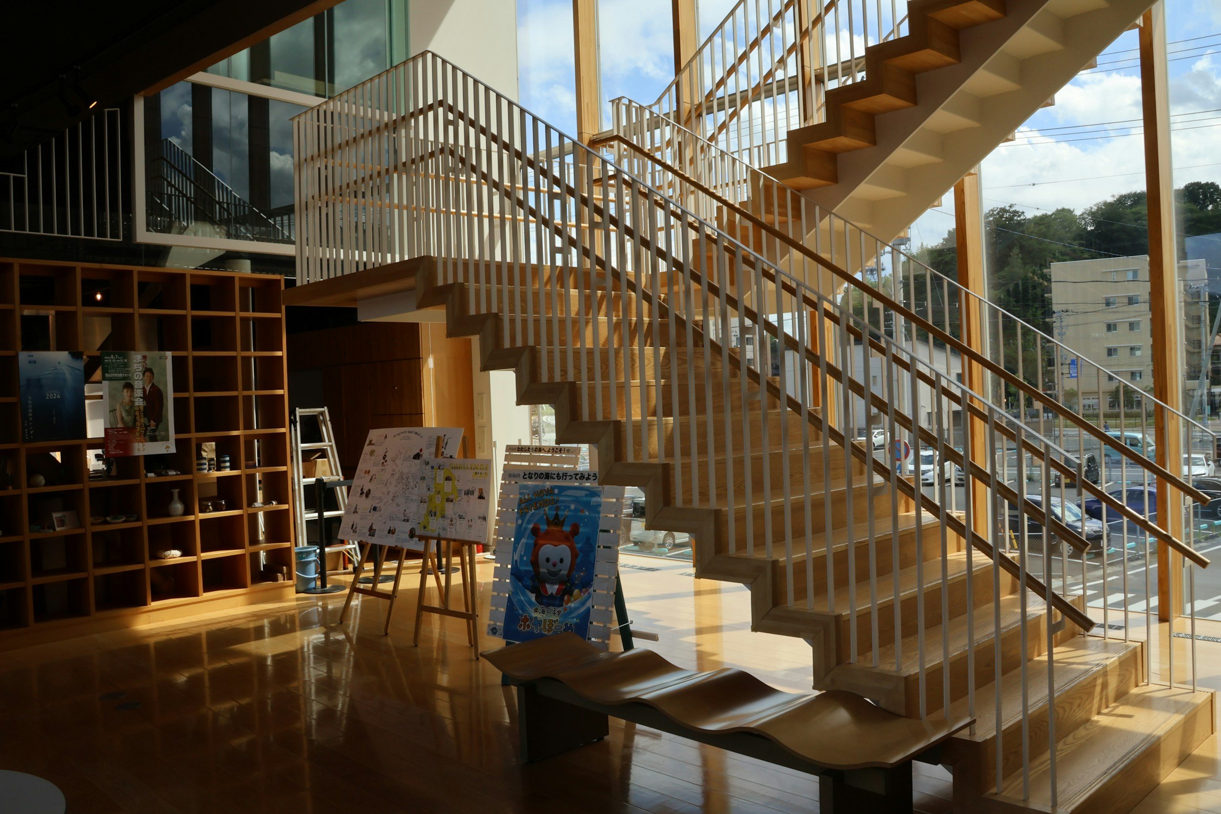 Interior of a modern building with a wooden staircase and large glass windows showing an outside parking lot and trees.