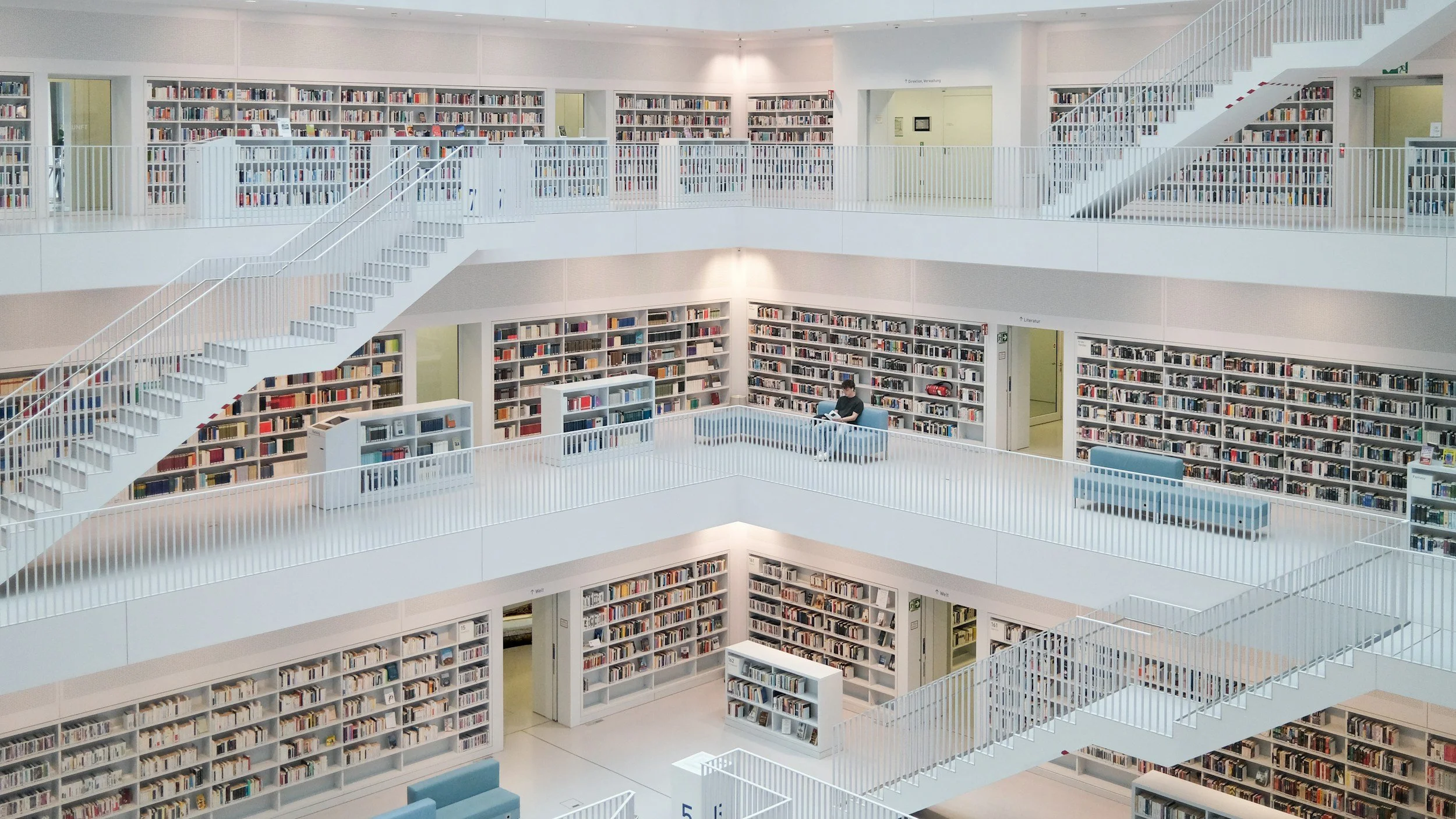 Interior of a modern multi-level library with white shelves filled with books, open spaces, and a staircase connecting the floors. A person is sitting and reading on the middle level.