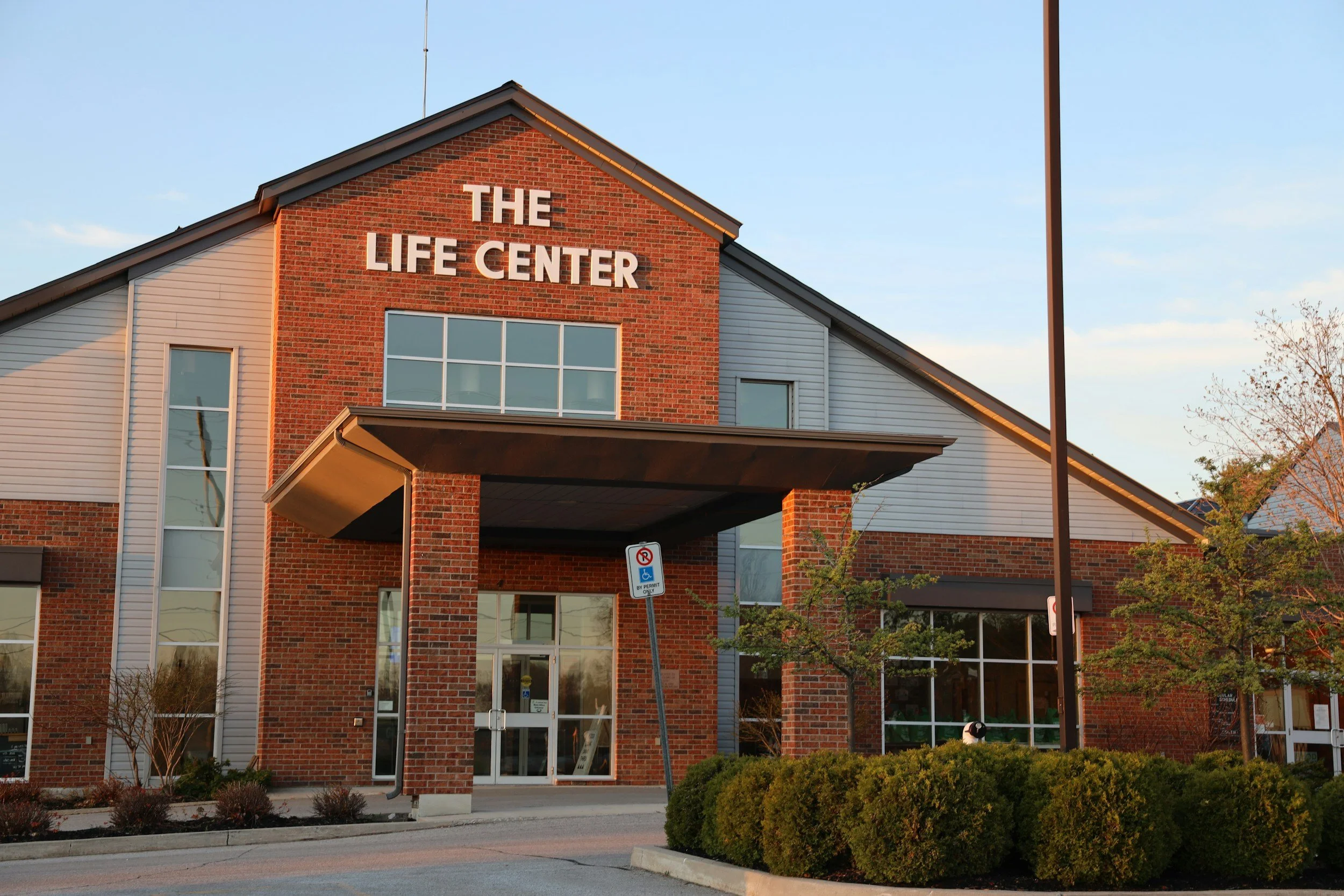 A brick and beige building with the sign 'The Life Center' on the front, surrounded by trees and bushes, with a parking sign in front.