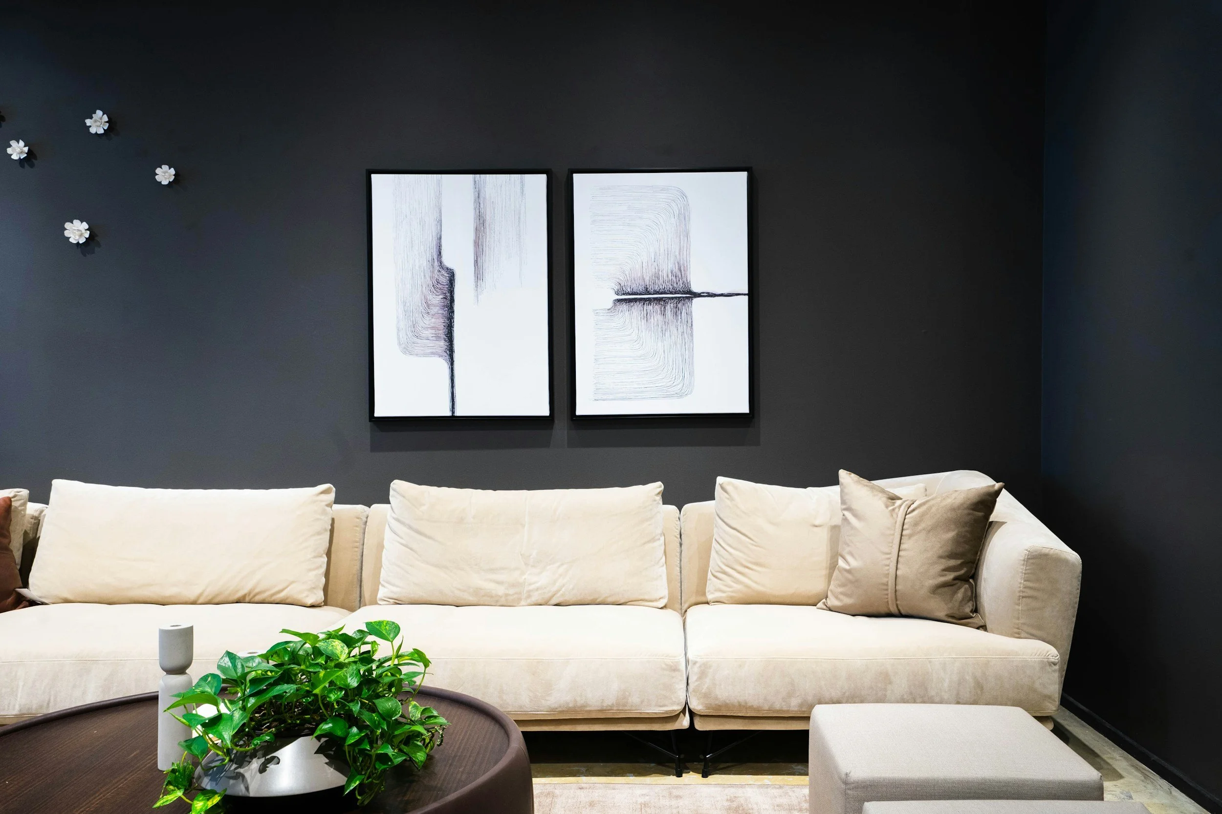 Modern living room with a white sofa, black wall, split abstract black and white artwork, a small round table with a green plant, and decorative flowers on the wall.