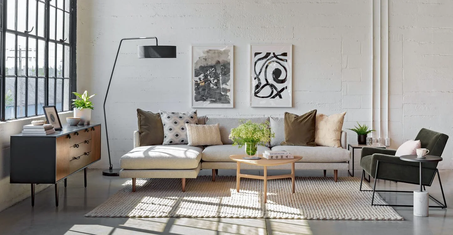 Modern living room with large window, white brick wall, beige sofa with patterned cushions, black floor lamp, wooden sideboard with books and decorative items, black and white abstract art, dark green armchair, small side table with plant, beige rug, and coffee table with flowers.