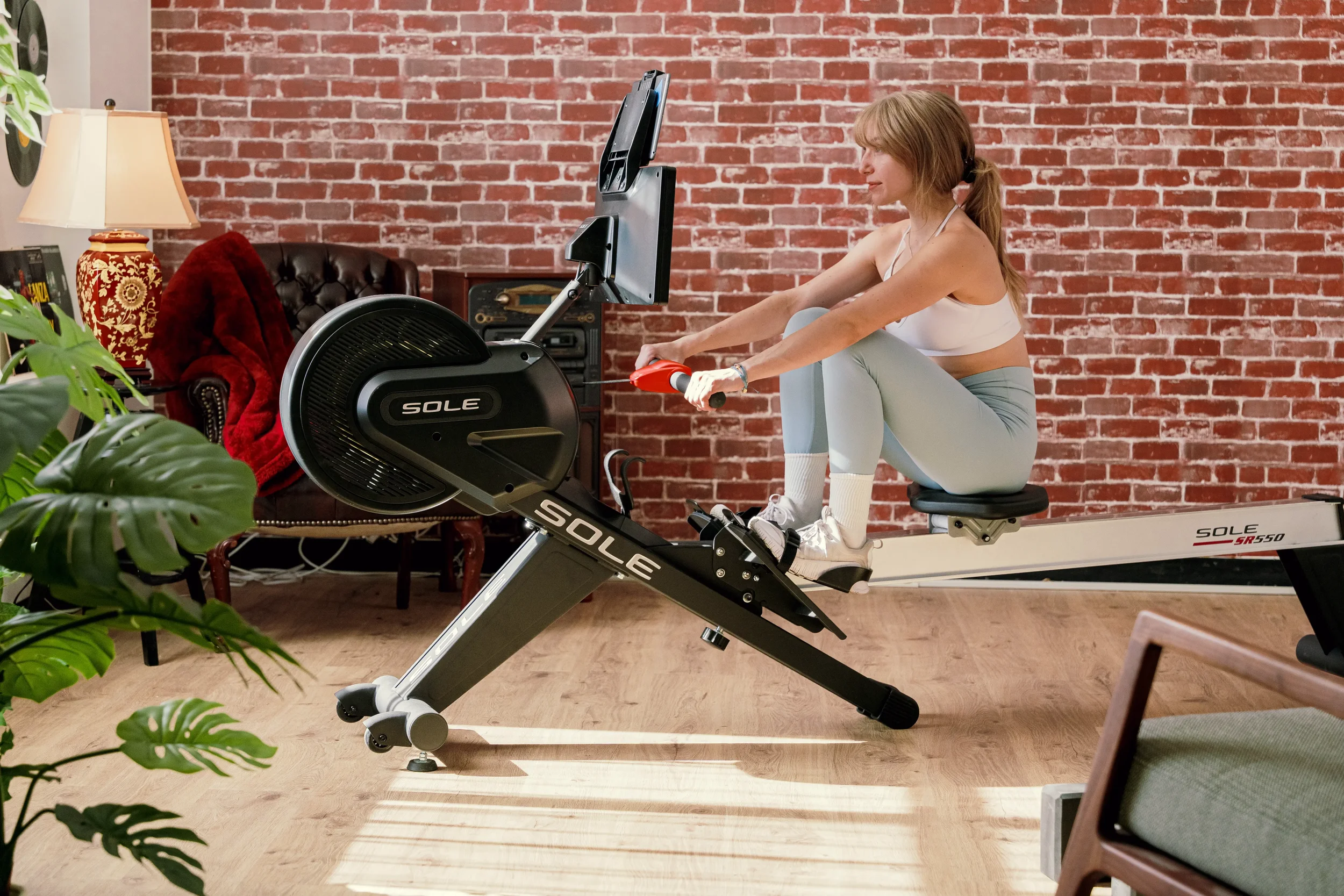 A woman in workout clothes using a rowing machine in a room with a brick wall and wooden floor, with furniture and a lamp visible in the background.