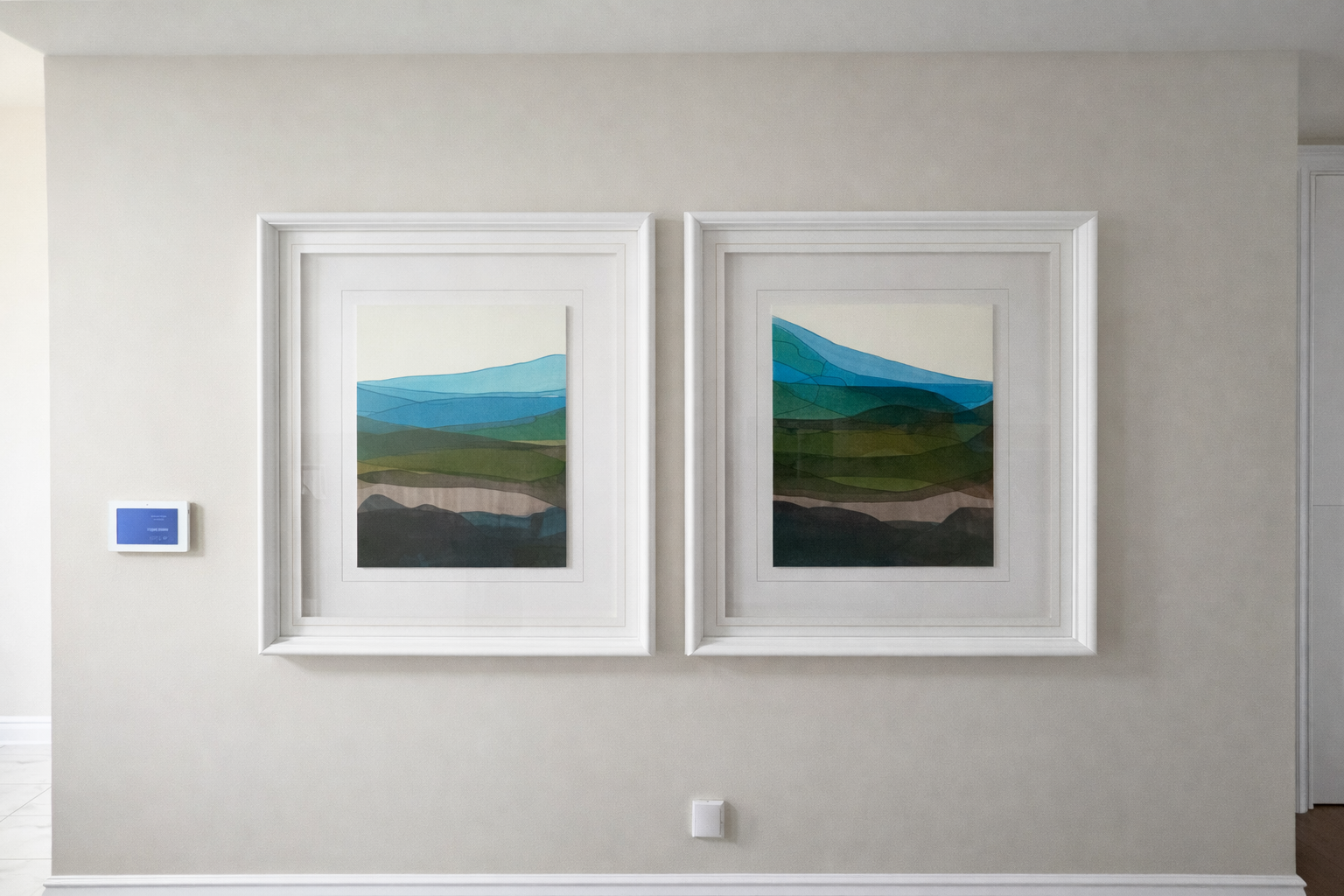 Two framed landscape paintings of mountains and hills hang on a beige wall in a gallery or home setting.