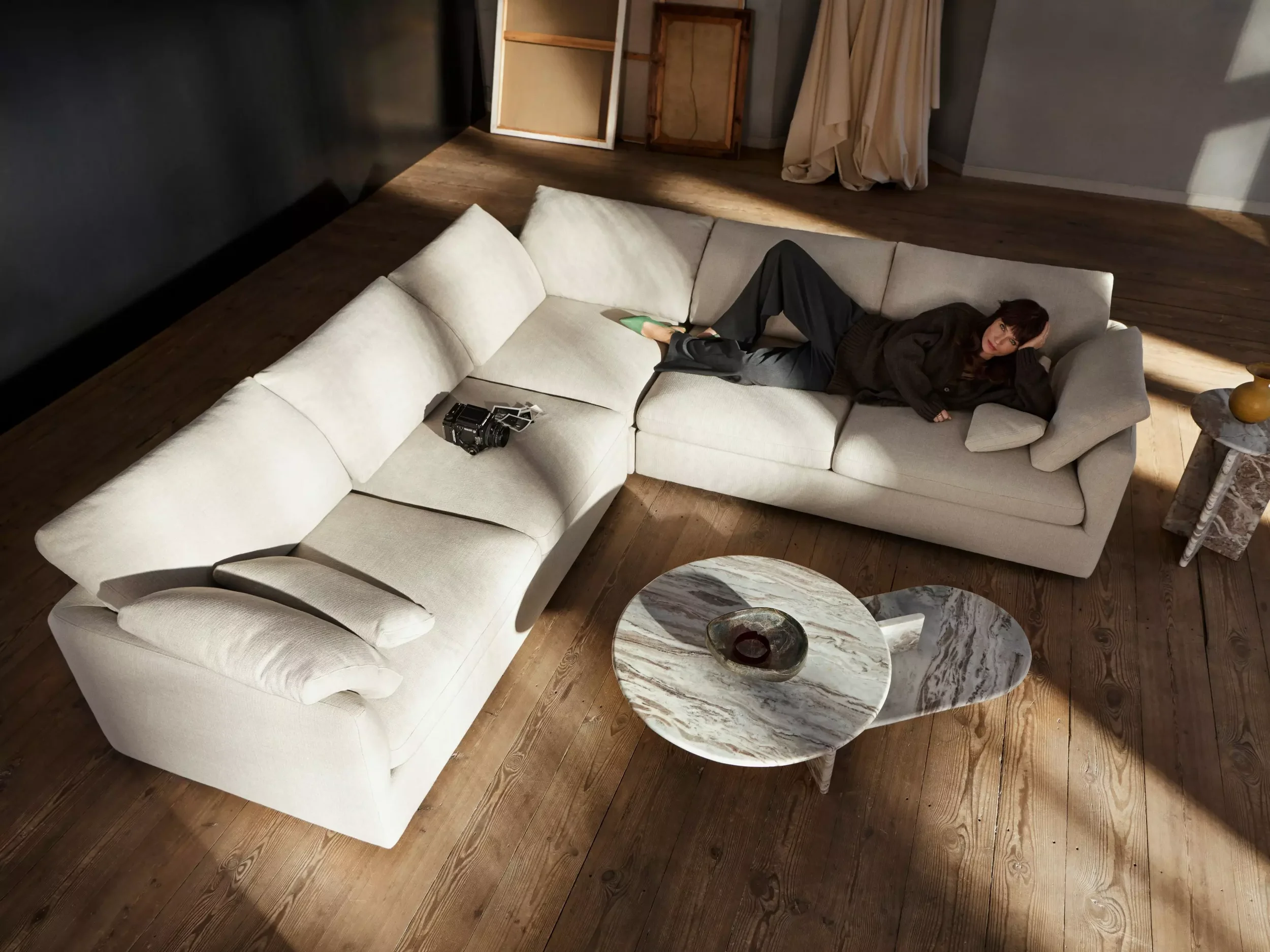 A woman in black clothing lying on a beige sectional sofa with a dark wall behind her, wooden flooring, and a marble coffee table in front with a small decorative bowl on top.