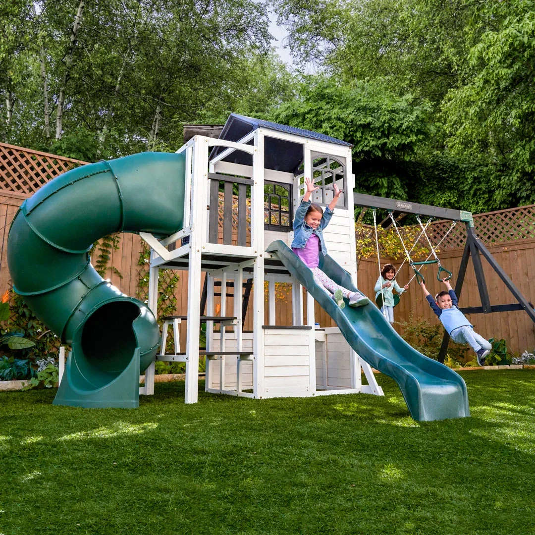 Children playing on a backyard playset with a slide, swings, and a enclosed playhouse surrounded by a wooden fence and green trees.