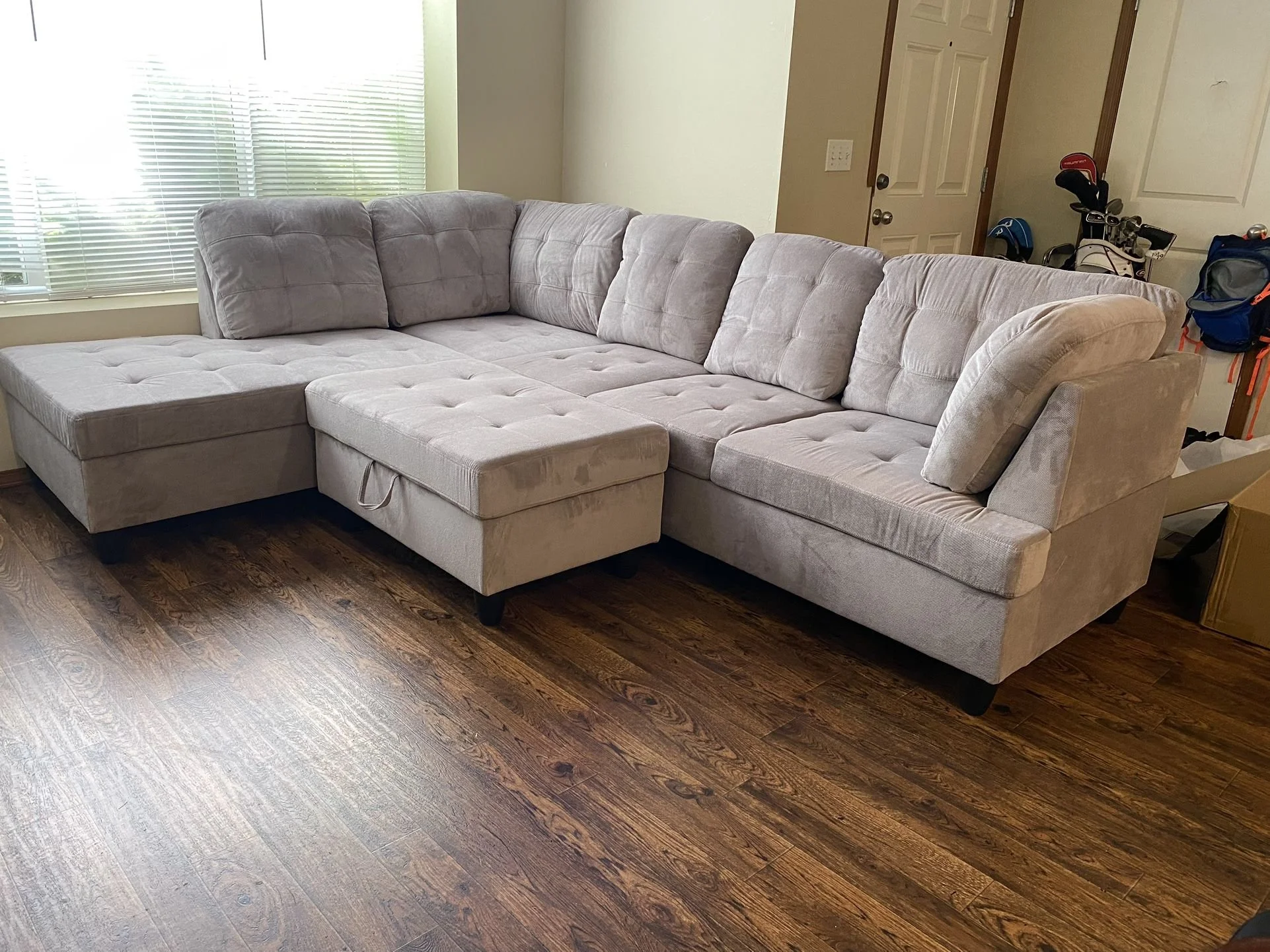 A light gray sectional sofa with a chaise lounge on the left side, positioned on a dark wood floor in a living room. Behind the sofa, there is a window with blinds and a nearby door on the right side. There are some golf clubs and bags near the door.