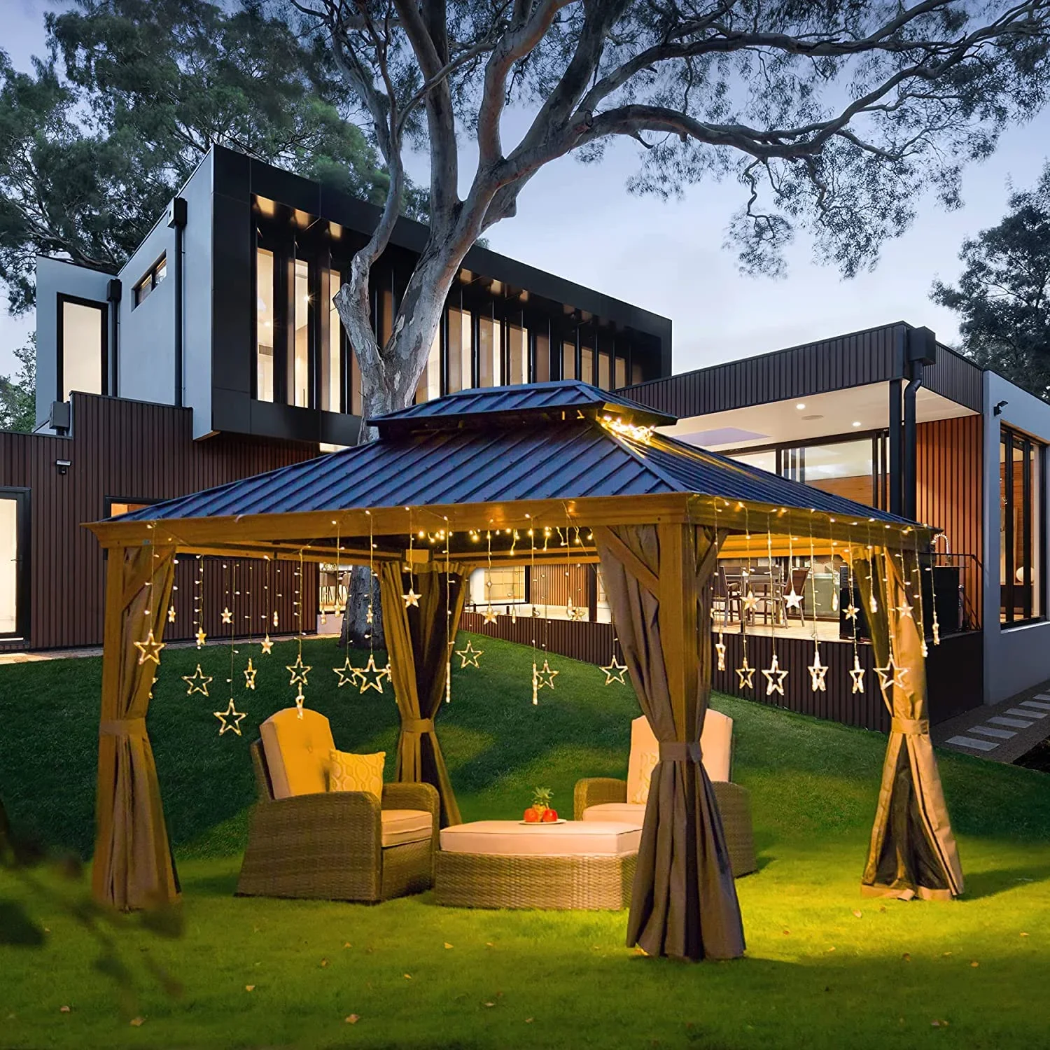 A cozy outdoor sitting area with a gazebo decorated with star-shaped string lights, set on a well-maintained lawn in front of a modern two-story house at dusk.