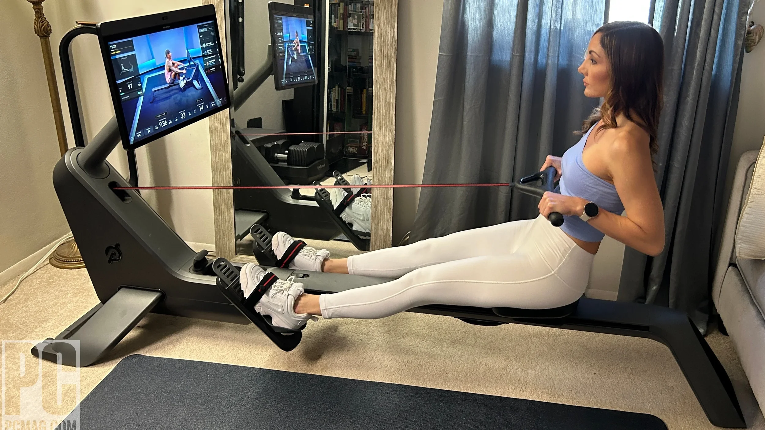 Woman exercising on a rowing machine, watching a workout video on a monitor in a home gym.