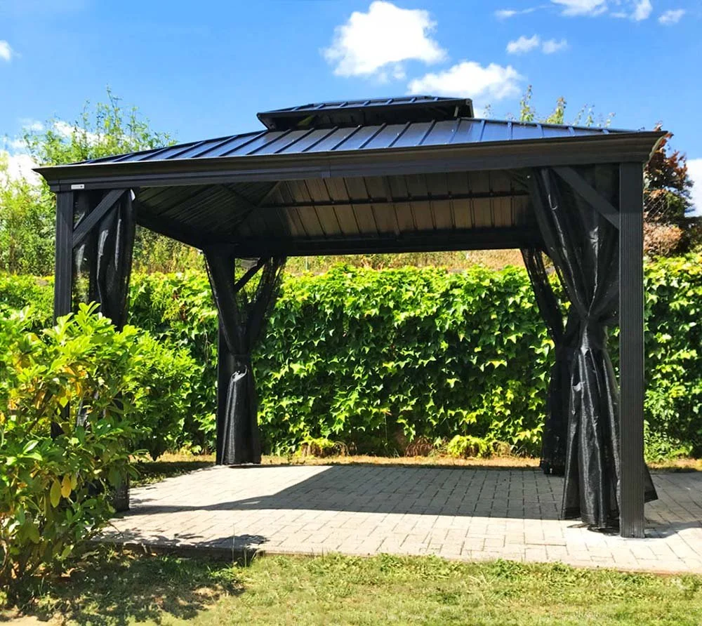 Black gazebo with curtains on a brick patio, surrounded by greenery under a partly cloudy sky.