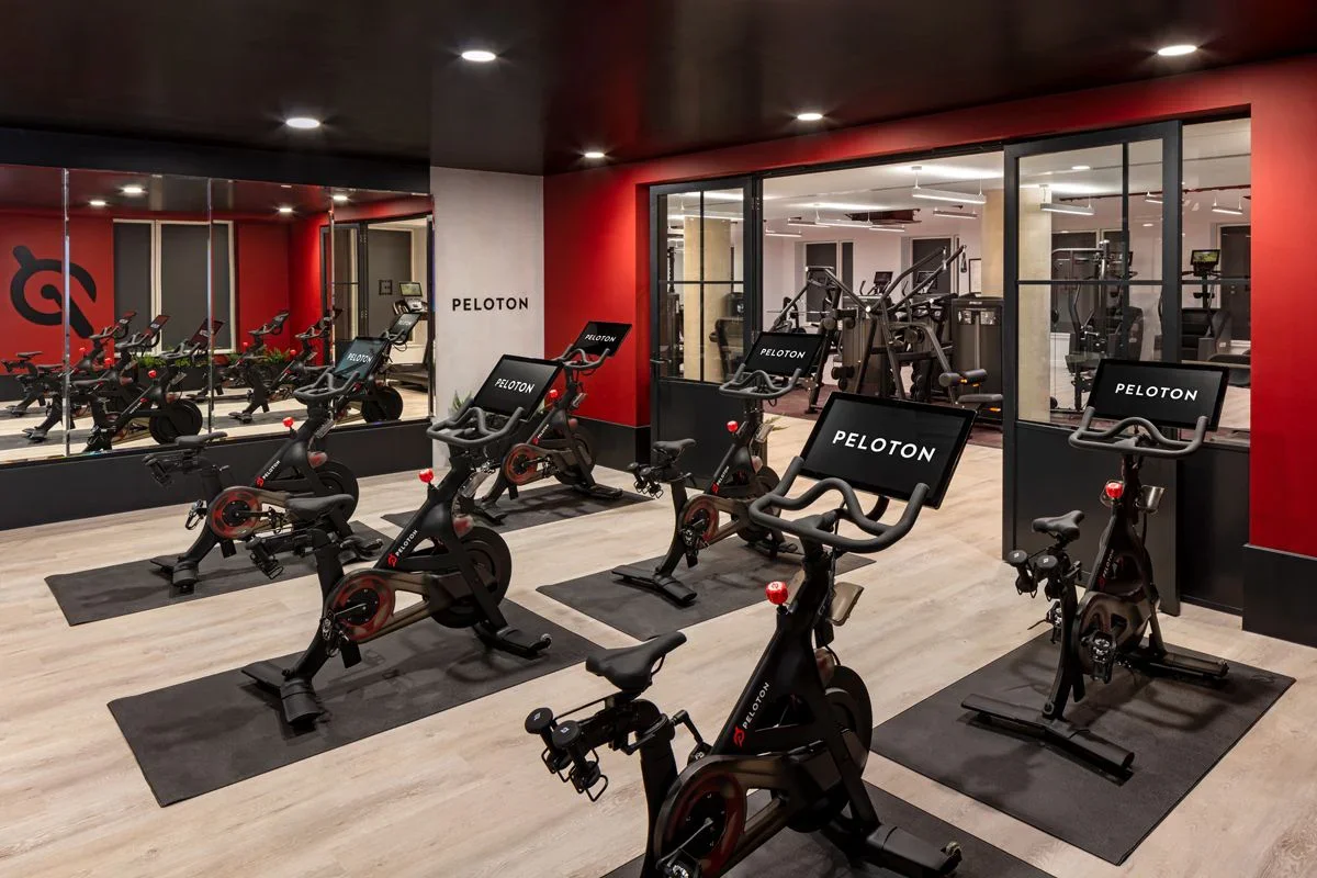 Indoor gym with Peloton stationary bikes arranged in rows on black mats, a large mirror on the left wall, and a glass wall separating the cardio area from the weight room, which contains various exercise machines.