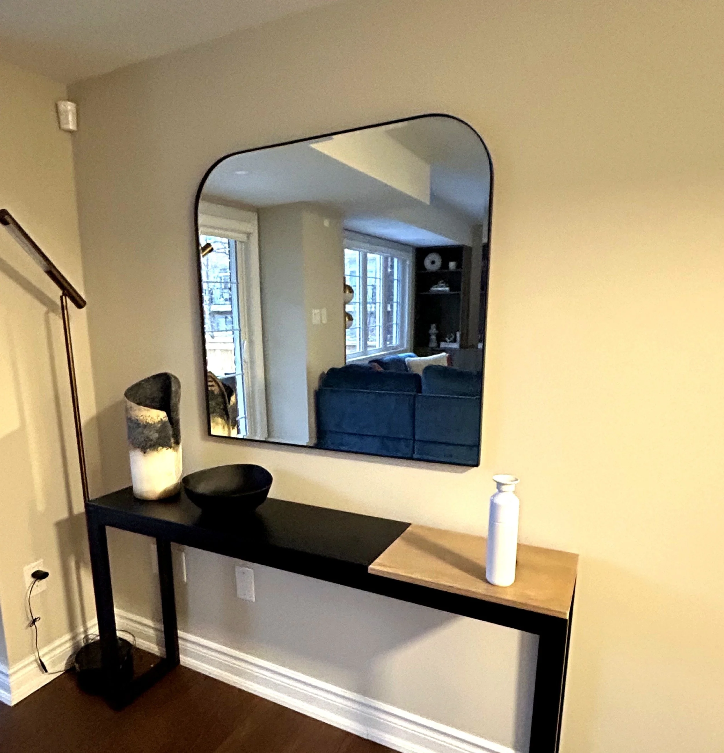 A modern console table with decorative vases, a bowl, and a white bottle is positioned against a beige wall, with a large rounded mirror reflecting a living room with a blue sofa, windows, and a bookshelf.