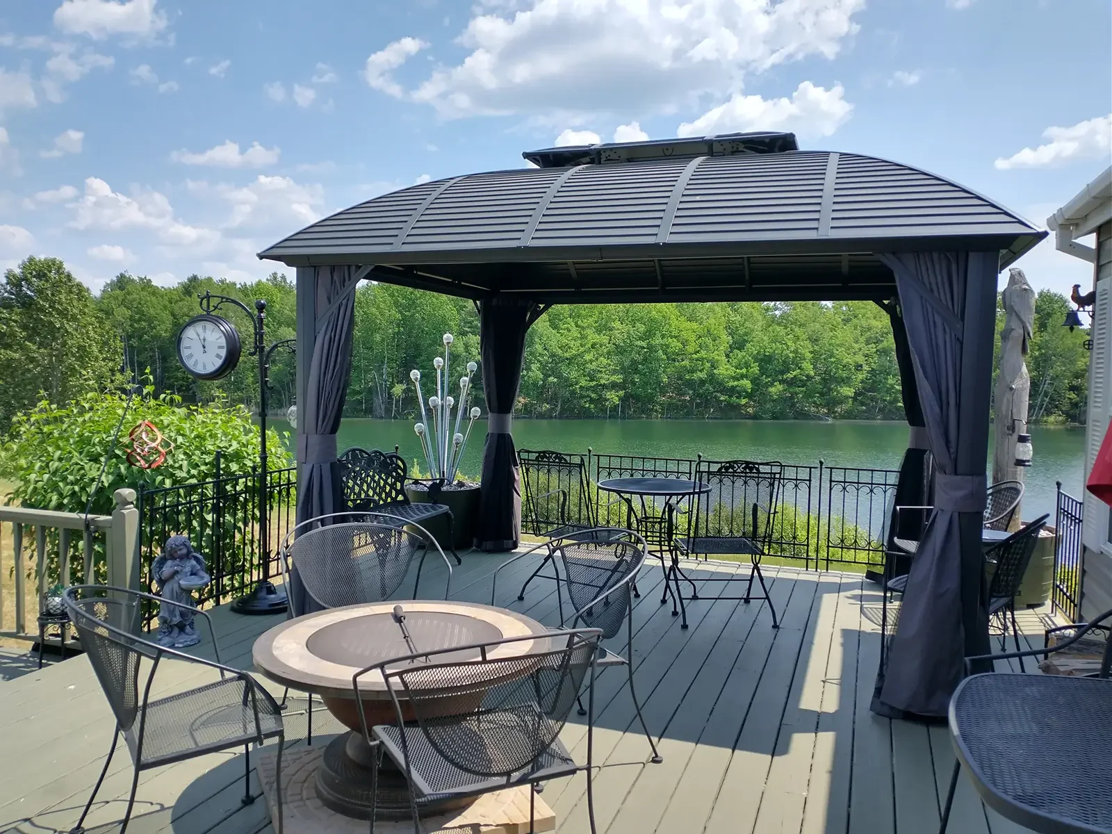 A lakeside patio with a black gazebo, outdoor furniture, and decorations, overlooking a lake with green trees in the background on a sunny day.