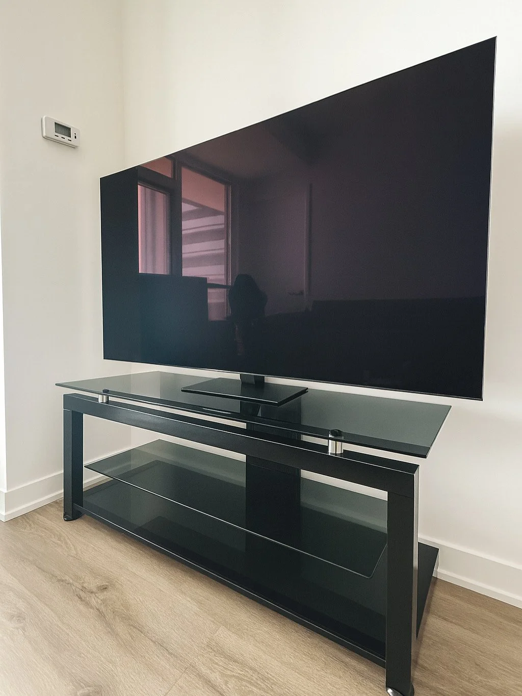 Large flat-screen TV on black glass stand in a room with white walls and wood flooring, with a reflection of windows and a shadowy figure visible on the TV screen.