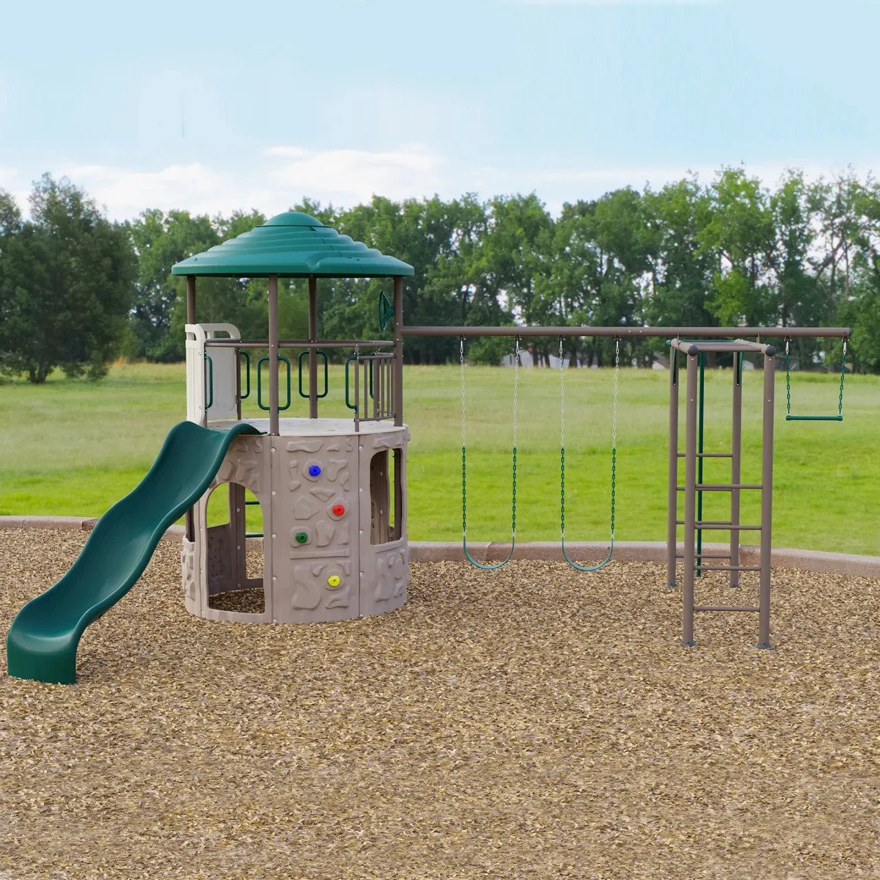 A playground with a slide, climbing wall, and swings on a gravel surface, with green grass and trees in the background.