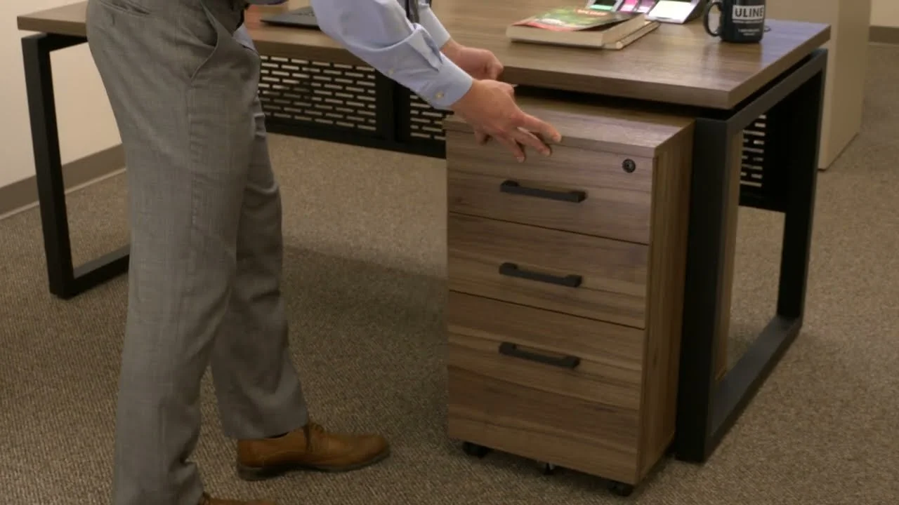 Person adjusting a small wooden filing cabinet with three drawers next to a desk in an office.