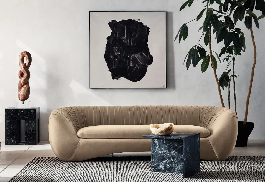 Modern living room with beige curved sofa, black marble side table with stone bowl, large potted plant, abstract black and white wall art, and a decorative sculpture on a black pedestal.