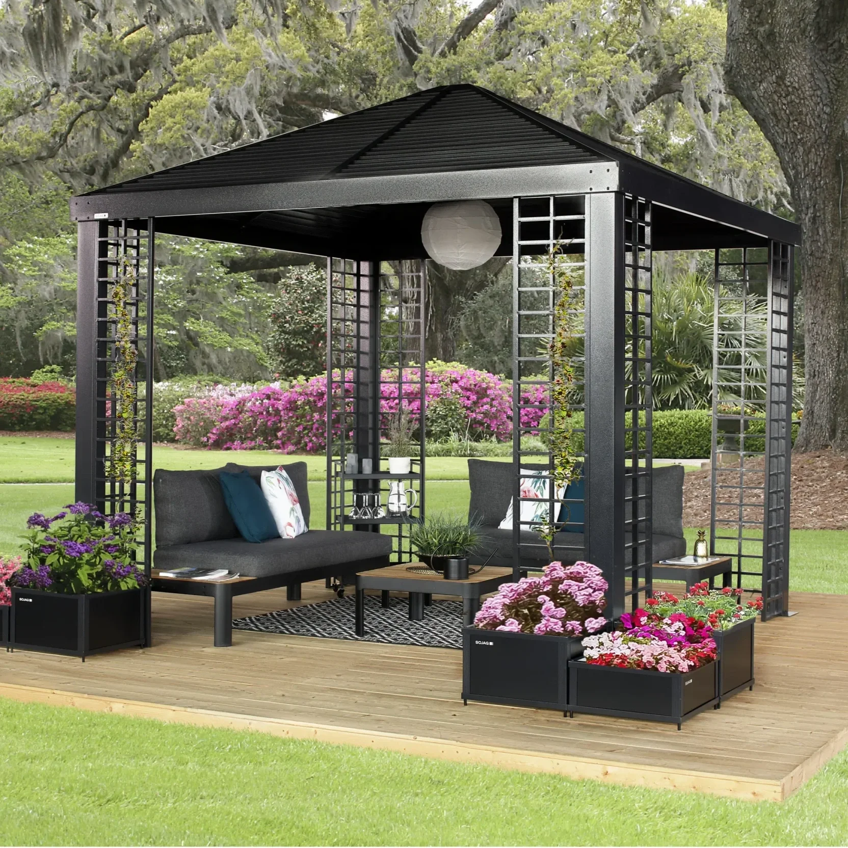A black outdoor pergola with seating, surrounded by potted flowers and plants, on a wooden deck in a garden with green grass and trees.