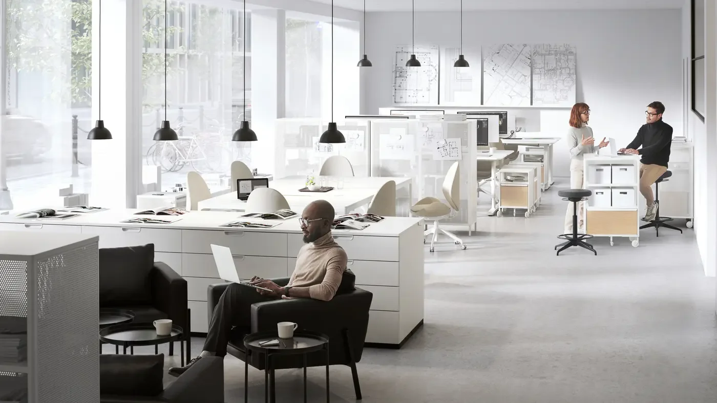 Modern open-plan office with large windows, white desks, ergonomic chairs, and a few employees working or talking, with plan images on the wall.