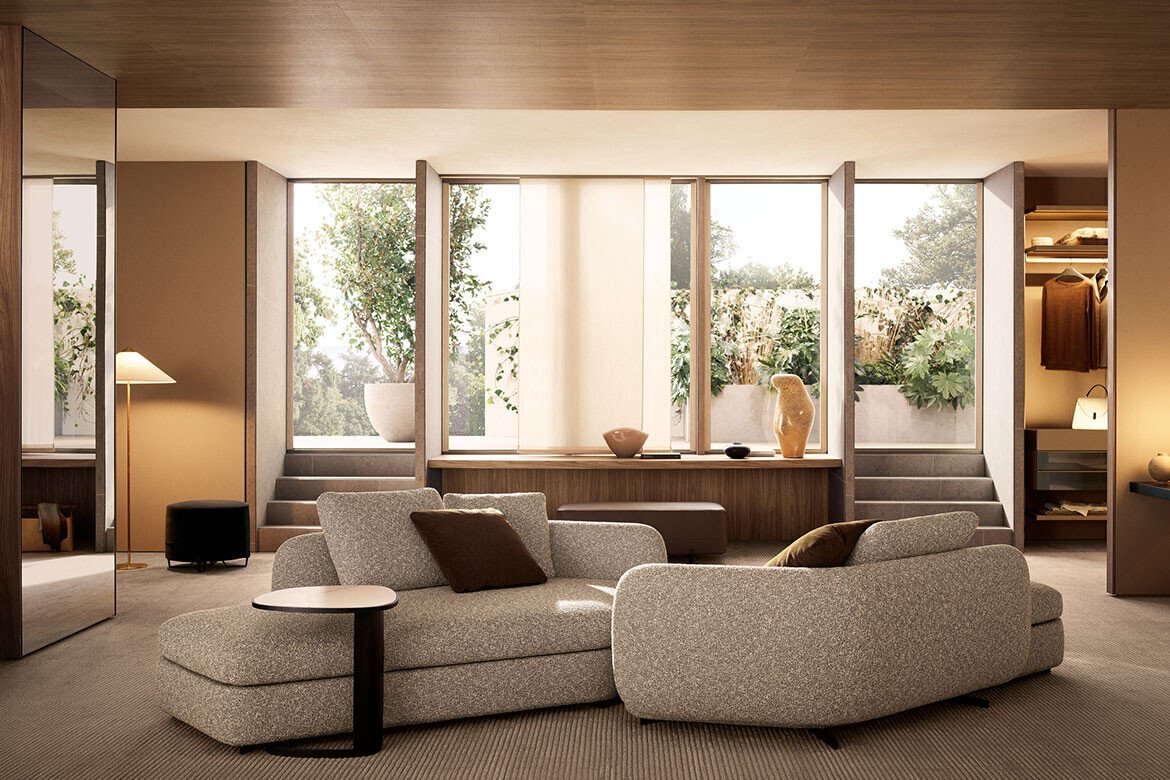 Living room with beige sofa, brown cushions, floor lamp, large windows with plants outside, wooden accents, and stairs leading to a higher level.