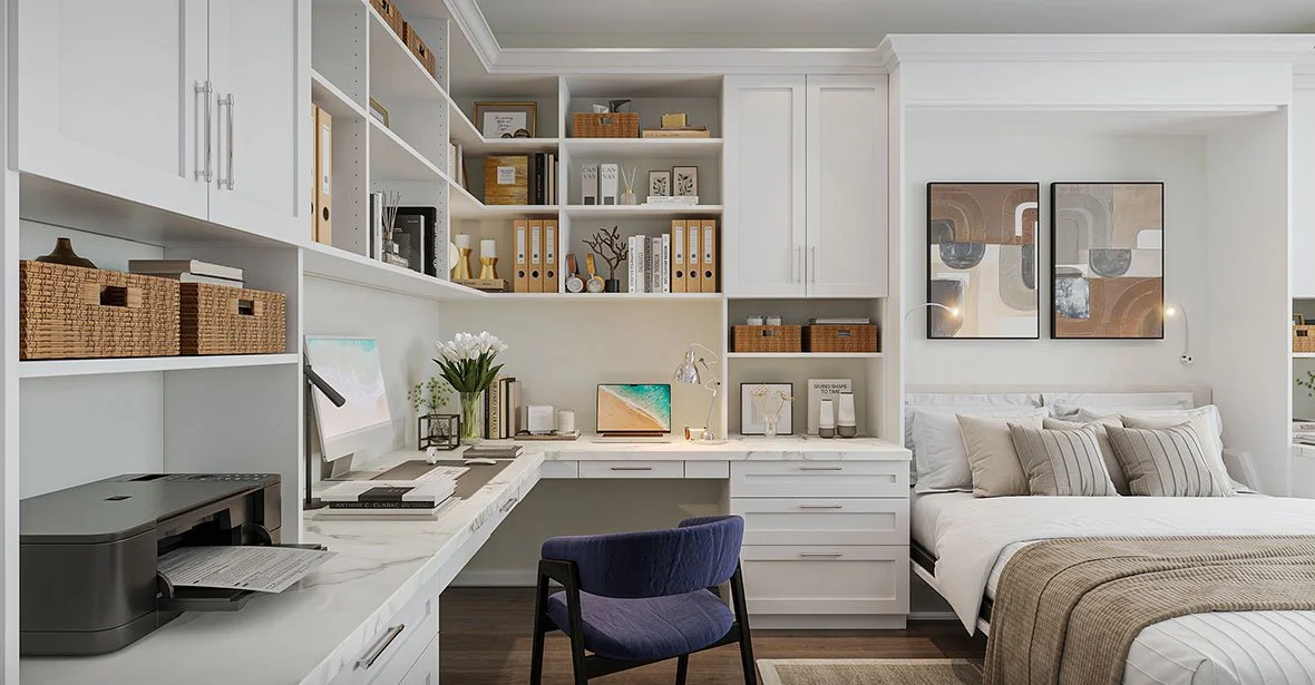 A bedroom with a built-in white desk and shelving, a computer monitor, a printer, a chair, a bed with pillows, and framed artwork on the wall.