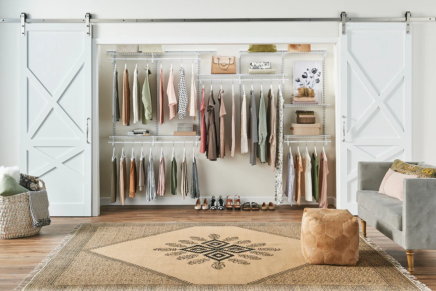 A neatly organized walk-in closet with white sliding barn doors, displaying hanging clothes, shelves with folded items, bags, and shoes, with a beige rug on wooden floor, a gray armchair, and a woven basket with pillows.