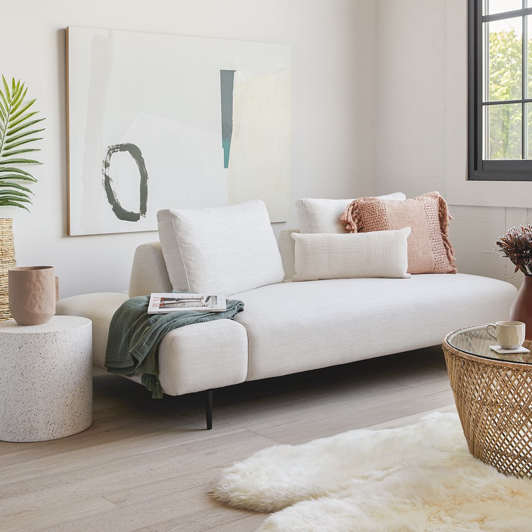 A cozy, minimalist living room with a white sofa decorated with assorted pillows, a pink throw blanket, and a side table with a magazine, a ceramic cup, and a pink vase. There's a large abstract painting on the wall, a window with natural light, a green plant, and a textured white rug on light wooden flooring.