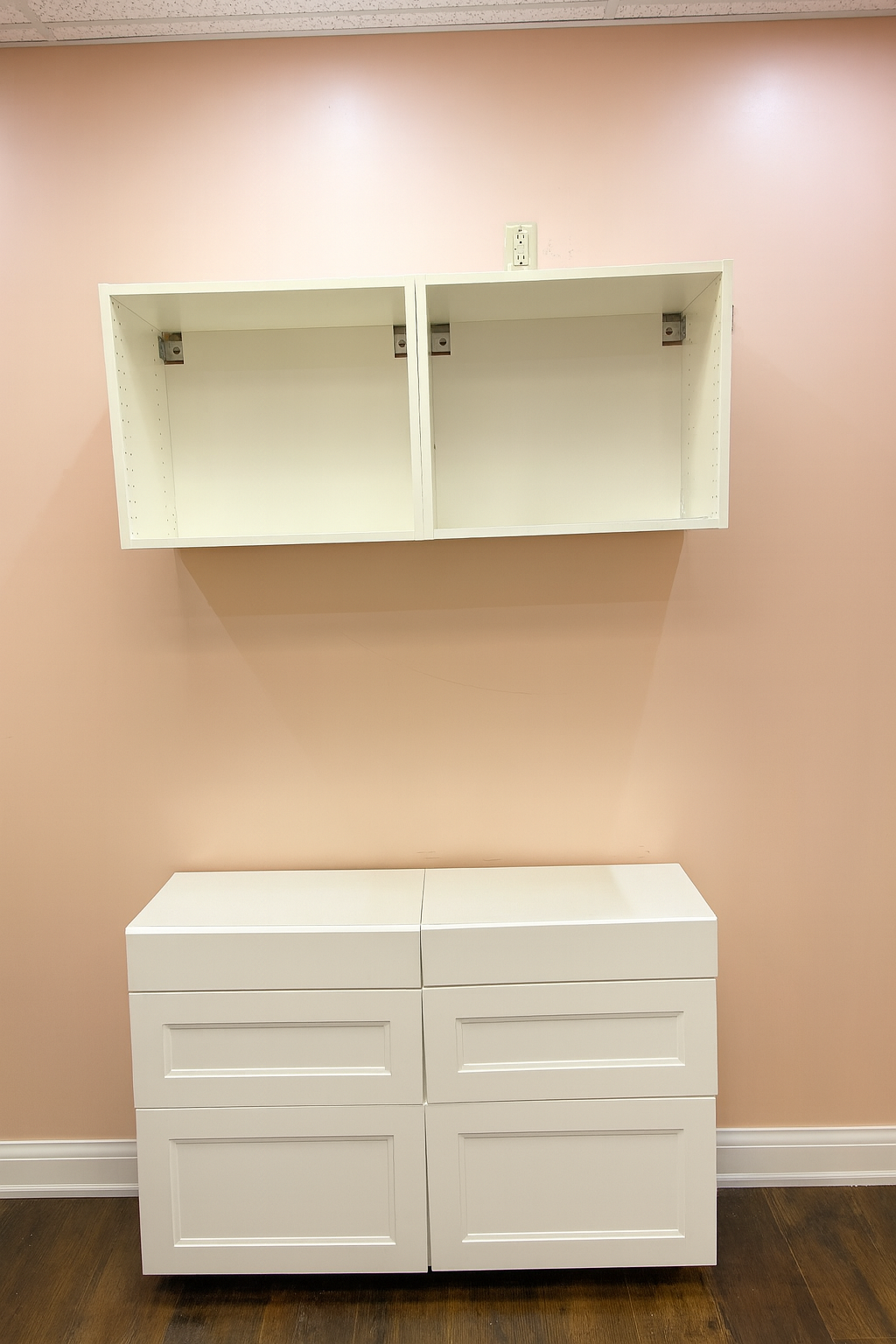 Empty white cabinet with two drawers below and an open shelf above, mounted on a pink wall with electrical outlets.
