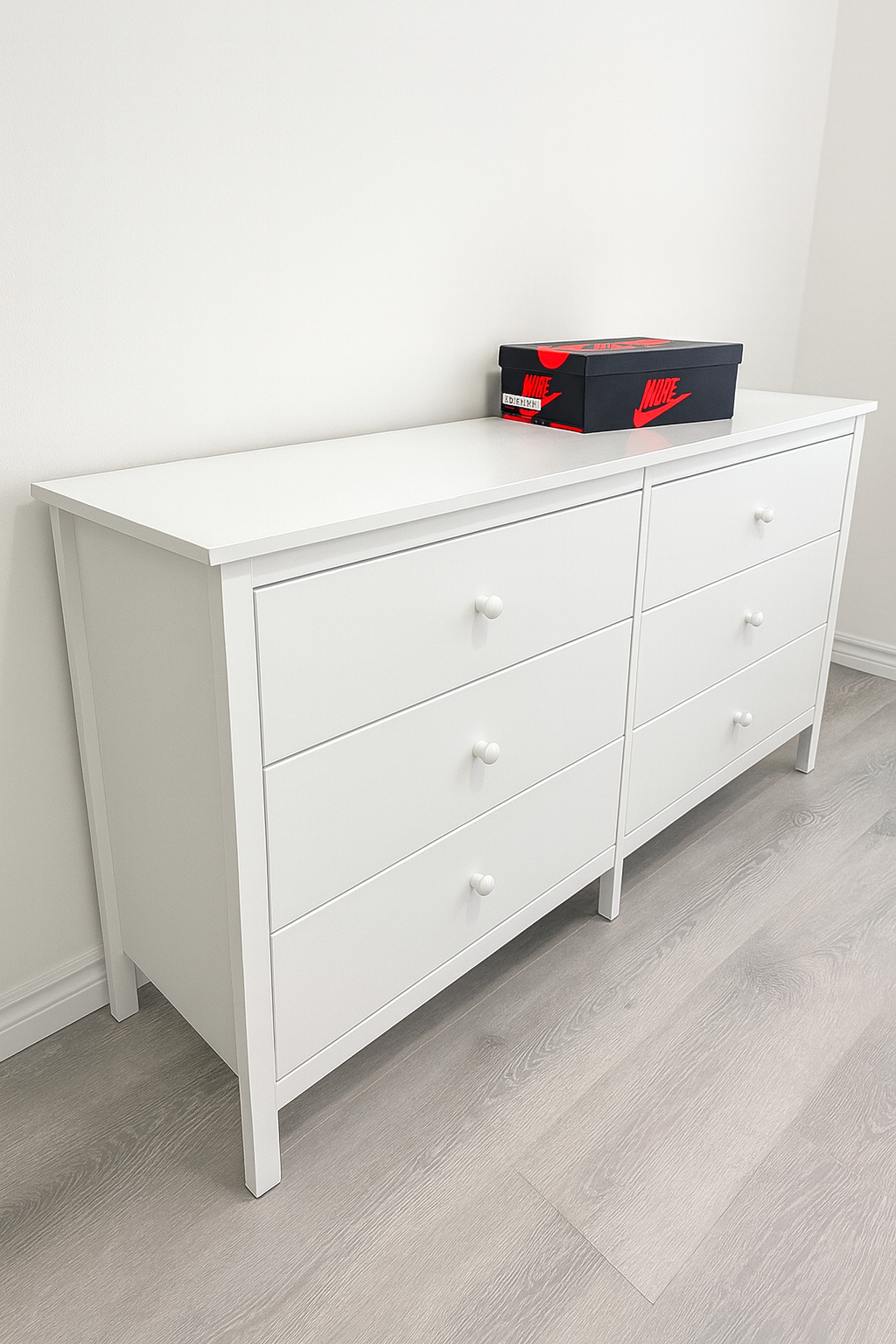 White dresser with six drawers and small round knobs, positioned against a white wall, with a black Nike shoe box on top.