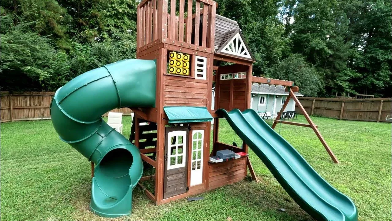 A wooden backyard playset with green slides, a tower, tic-tac-toe game, and a small clubhouse area, surrounded by a grassy yard and a wooden fence.