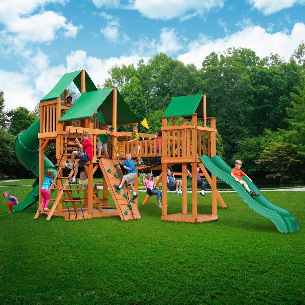 Children playing on a large wooden playground with green slides and a climbing wall in a park with green trees and blue sky.