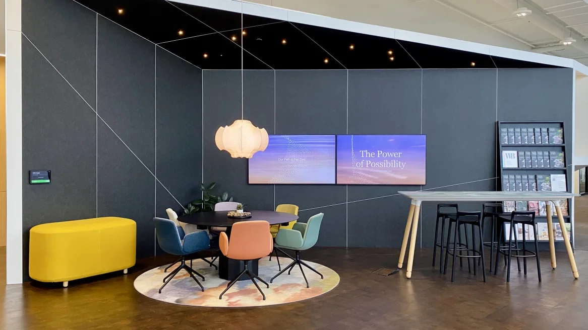 A modern lobby or lounge area with a round table surrounded by colorful chairs, a yellow bench, a hanging pendant light, a large digital screen displaying "The Power of Possibility," and magazines on a stand.