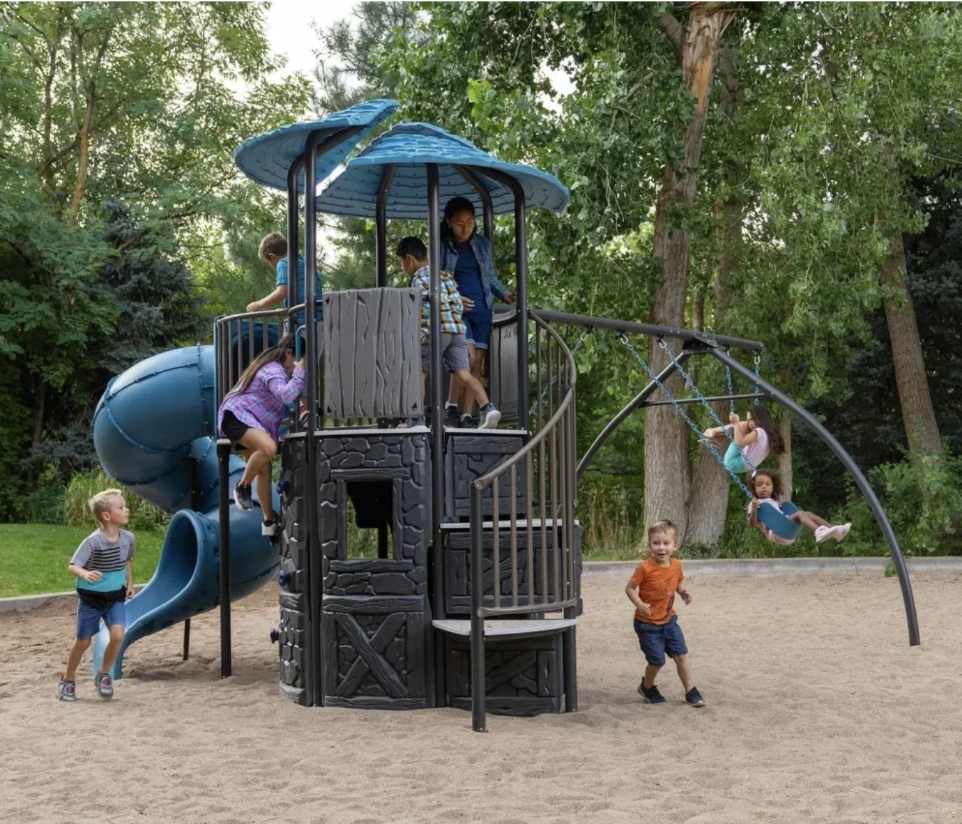 Children playing on a multi-level playground structure with a spiral slide and swings in a park surrounded by trees.