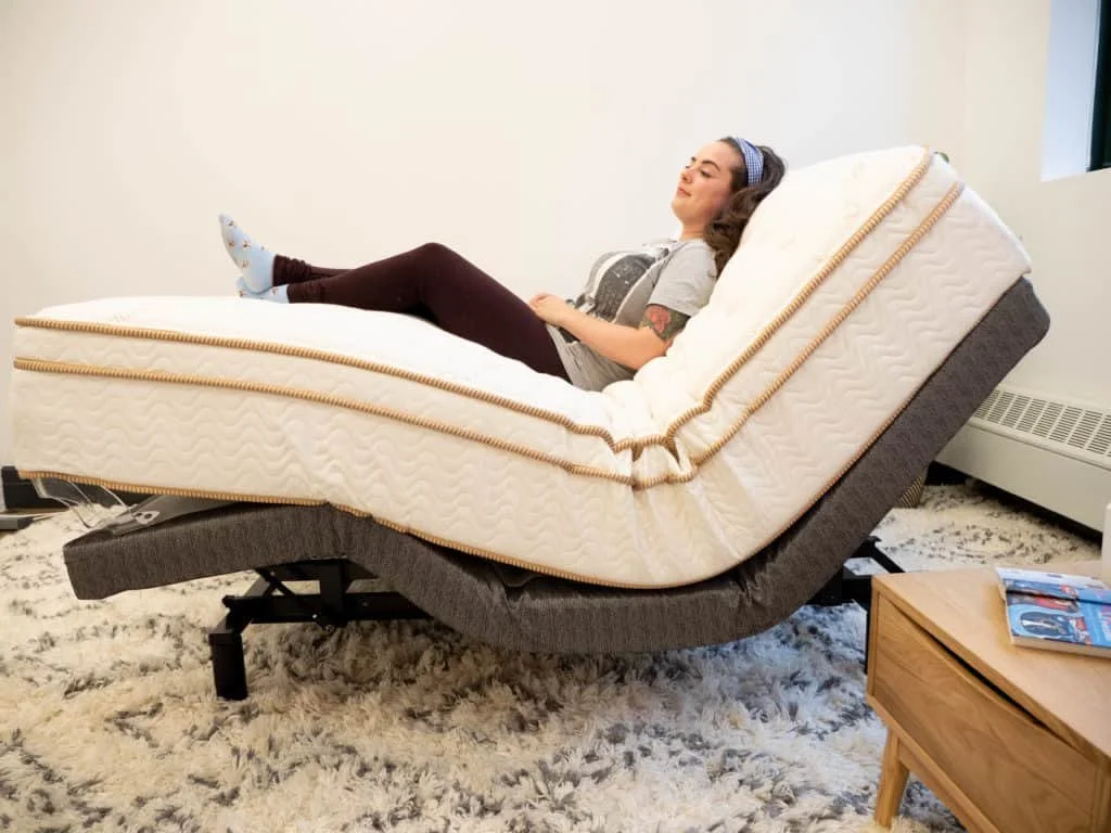 A woman relaxing on a tilt-up adjustable bed in a room with a beige carpet, a wooden side table, and a radiator.