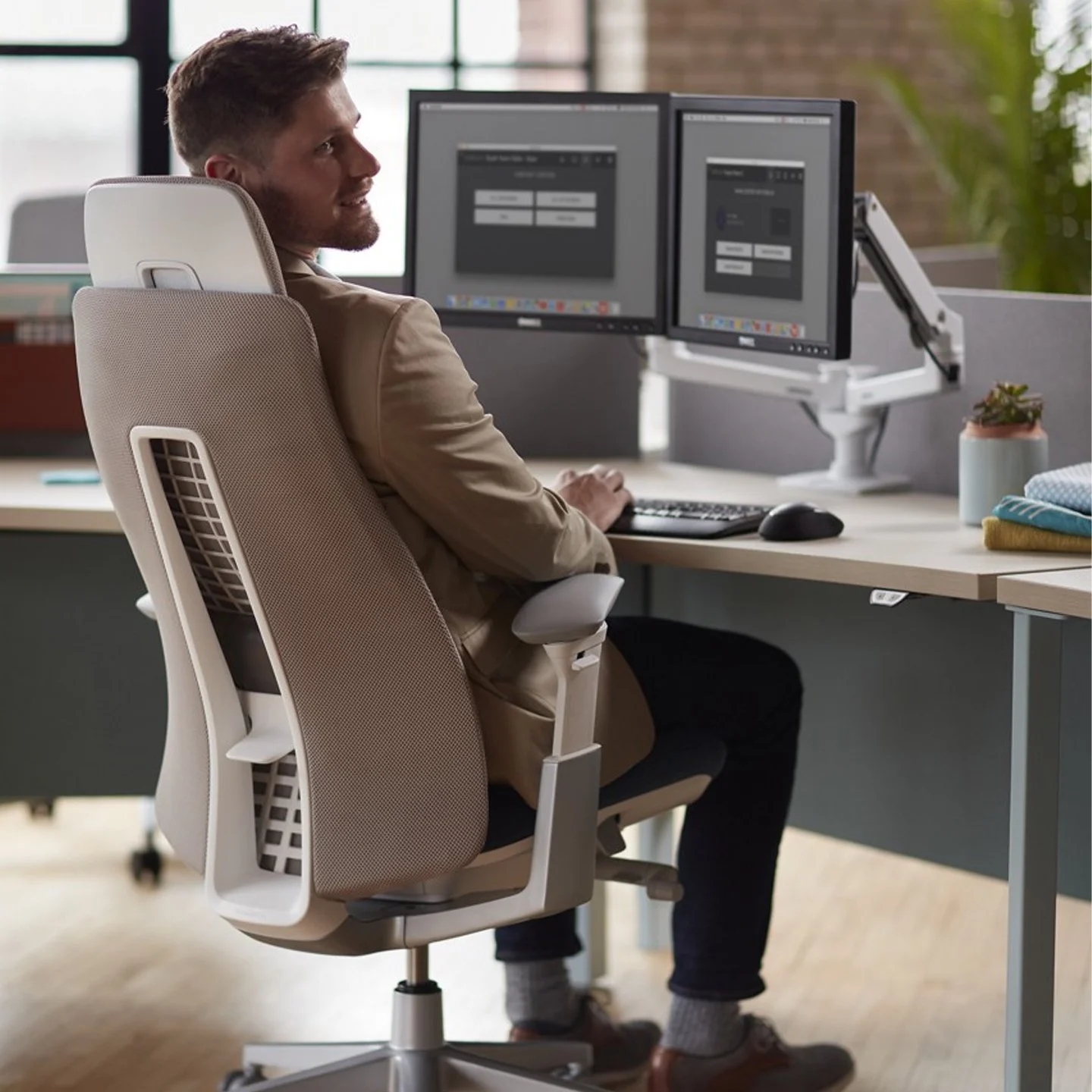 A man sitting at a desk with dual monitors in an office space, looking to his right with a smile.
