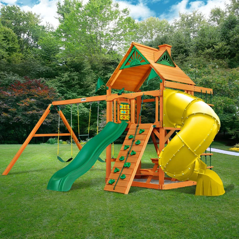 A wooden playground set with a slide, swings, a climbing wall, and a yellow tube tunnel, situated on a grassy area with trees in the background.