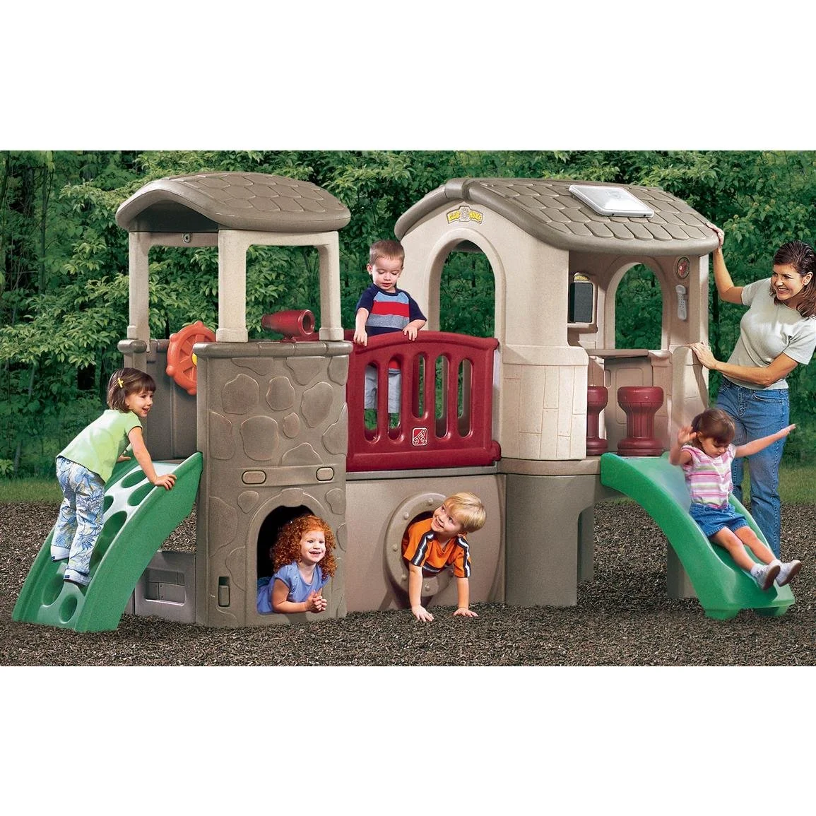 Children playing on a multi-level plastic playground structure outdoors with slides, a rock climbing wall, and a small house, supervised by an adult woman.