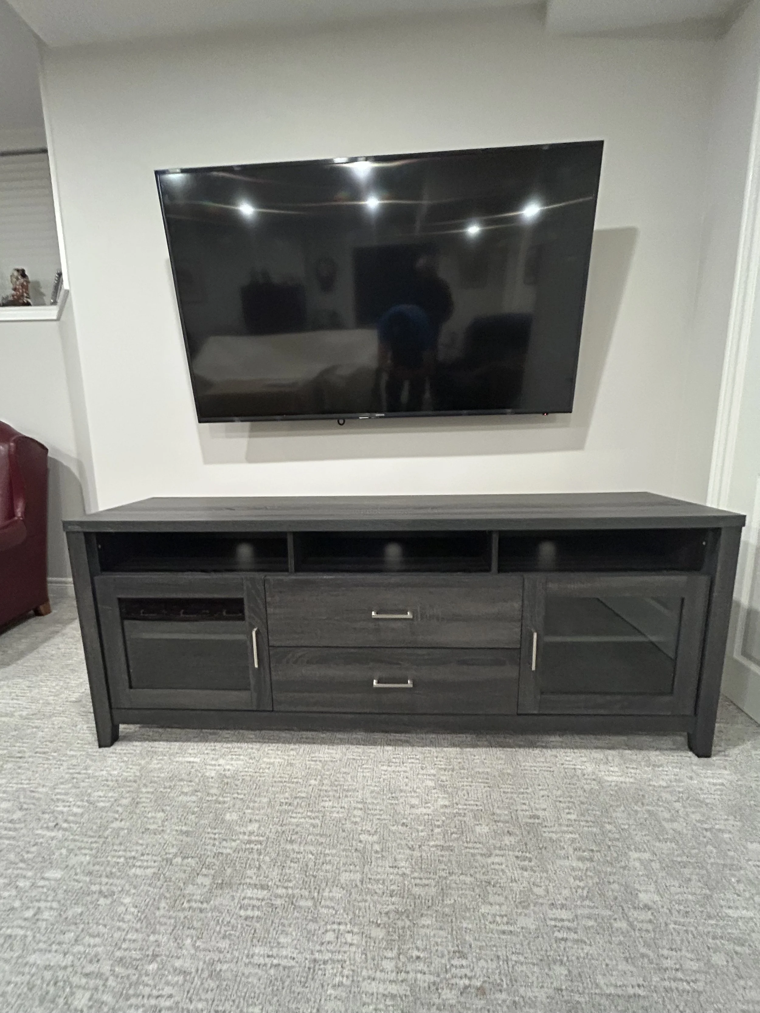 Black TV stand with open cubbies and drawers, with a large flat-screen TV mounted above. Reflection of person taking the photo visible on TV screen. Part of a red chair visible on the left, with beige carpeted floor.