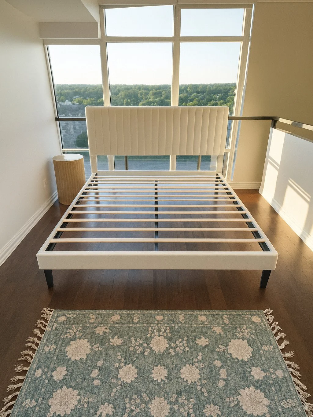 Unmade white bed frame with a high padded headboard in a high-rise bedroom, with large windows showing a green landscape outside, a round beige nightstand on the left, and a teal floral rug on a wooden floor.
