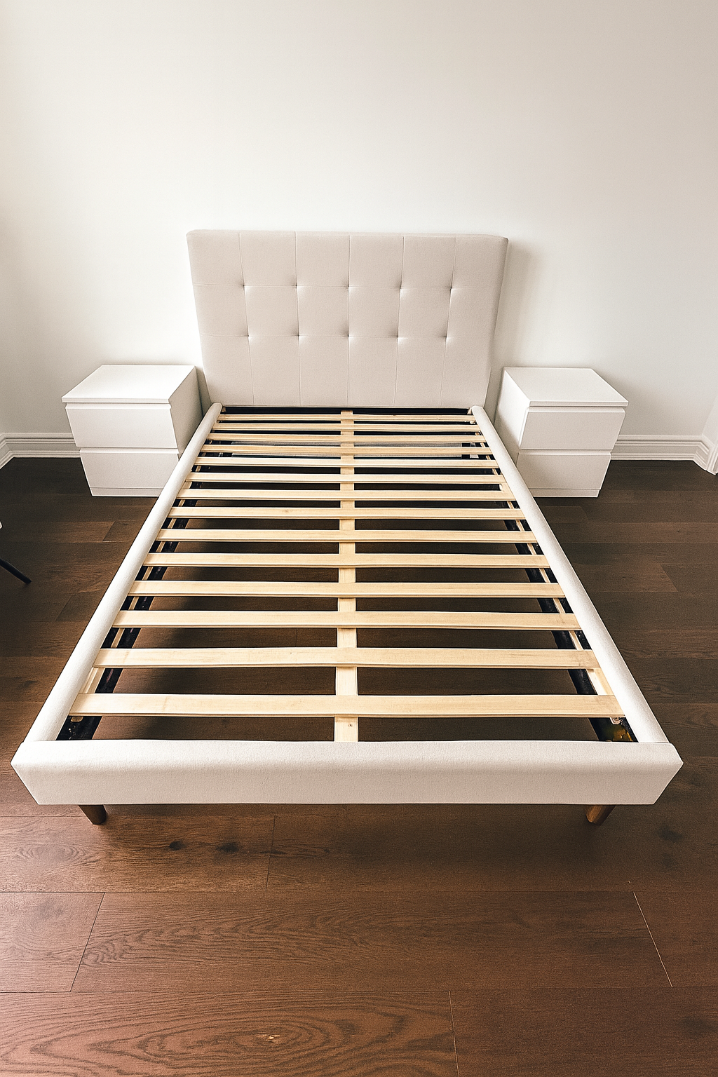 Empty bed frame with a tufted headboard and two white nightstands in a bedroom with dark hardwood floors.
