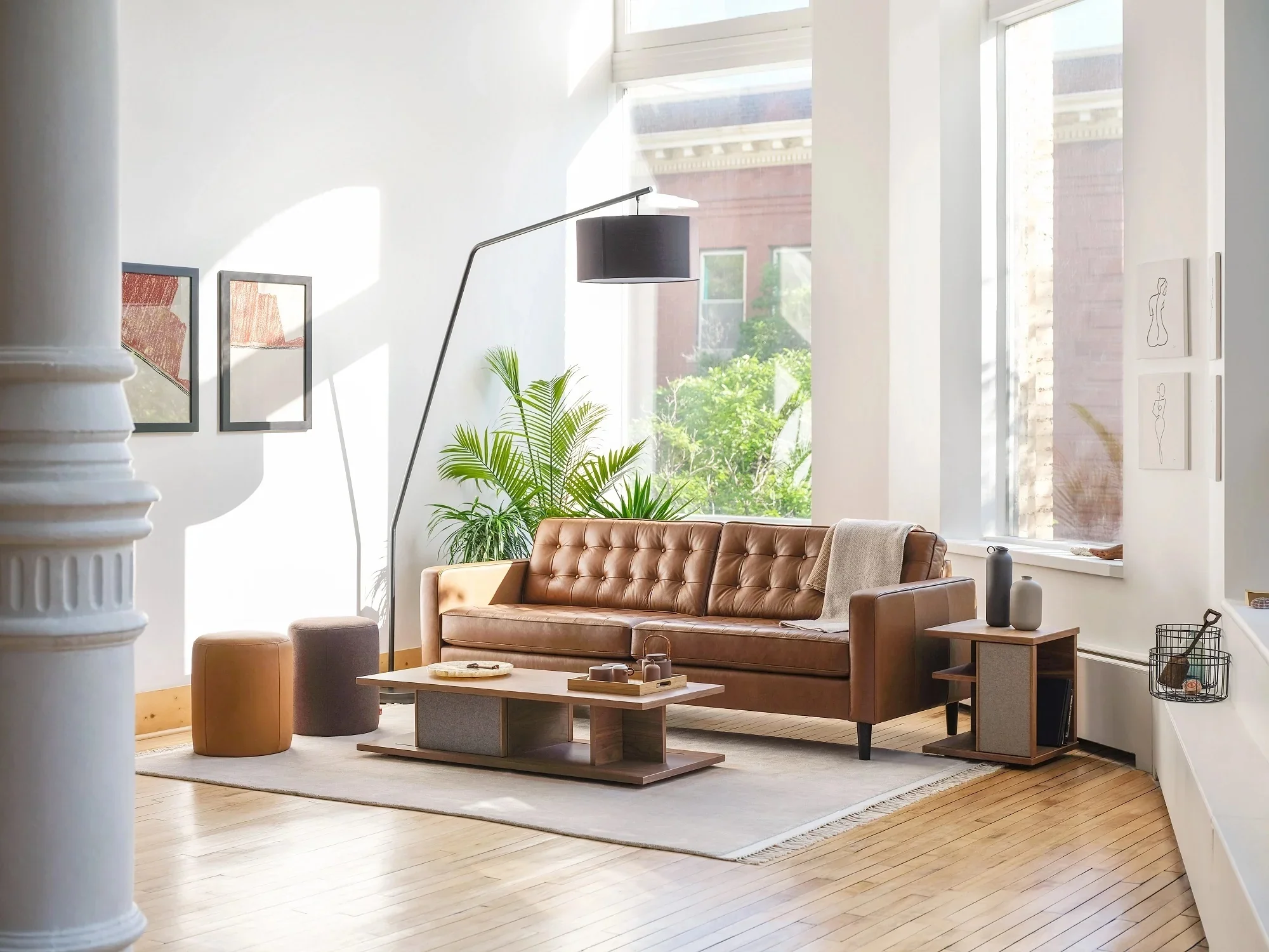 Modern living room with a brown leather sofa, wooden coffee table, tall floor lamp, large window with sunlight, and art pieces on the wall.