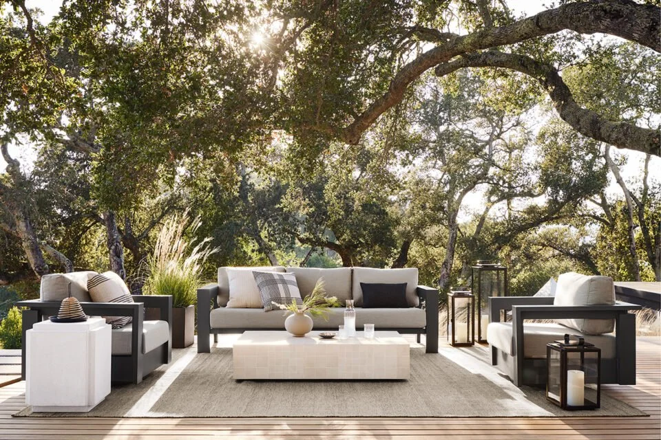 Outdoor patio with modern white and black seating, including a sofa and armchairs, decorated with pillows, surrounded by greenery and trees, with sunlight filtering through