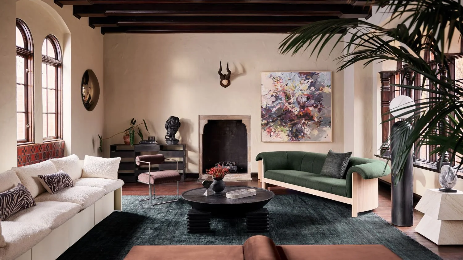A living room with beige walls, dark wood ceiling beams, and a fireplace. It has two sofas, one beige with zebra-patterned pillows and a green one with a dark pillow. There are large arched windows with wooden frames, plants, artwork, and a modern coffee table with decorative items in the center.