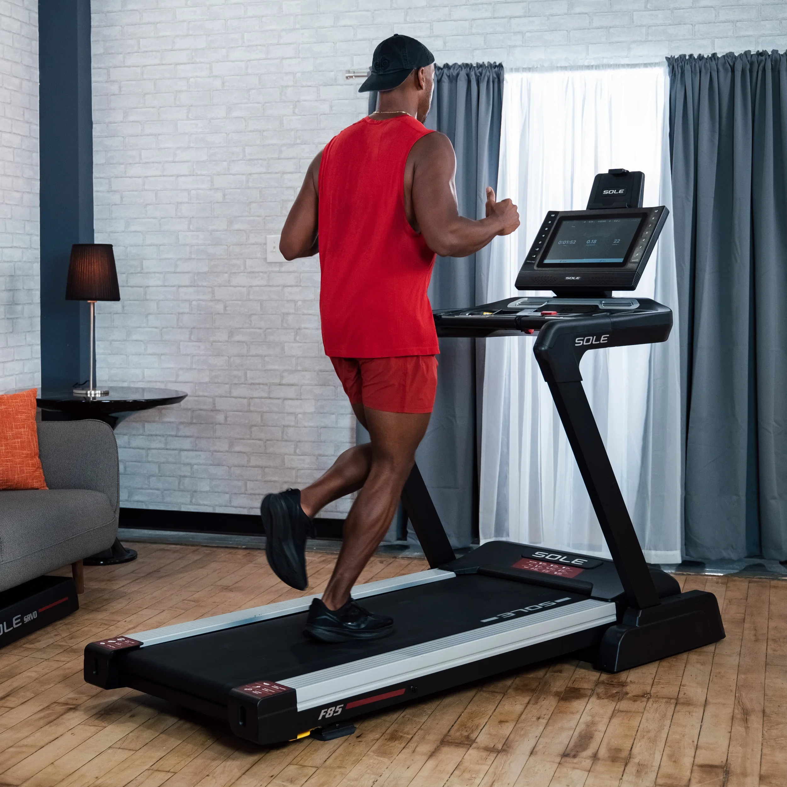 Man running on a treadmill in a home gym with curtains and a sofa in the background.