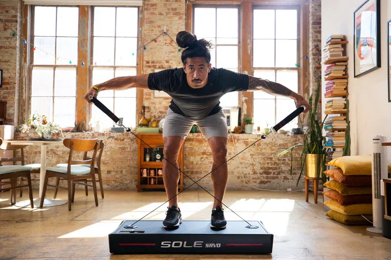 A man with a top knot hairstyle is exercising on a rebounder in a bright, industrial-style room with large windows, wooden chairs, and a stack of books.