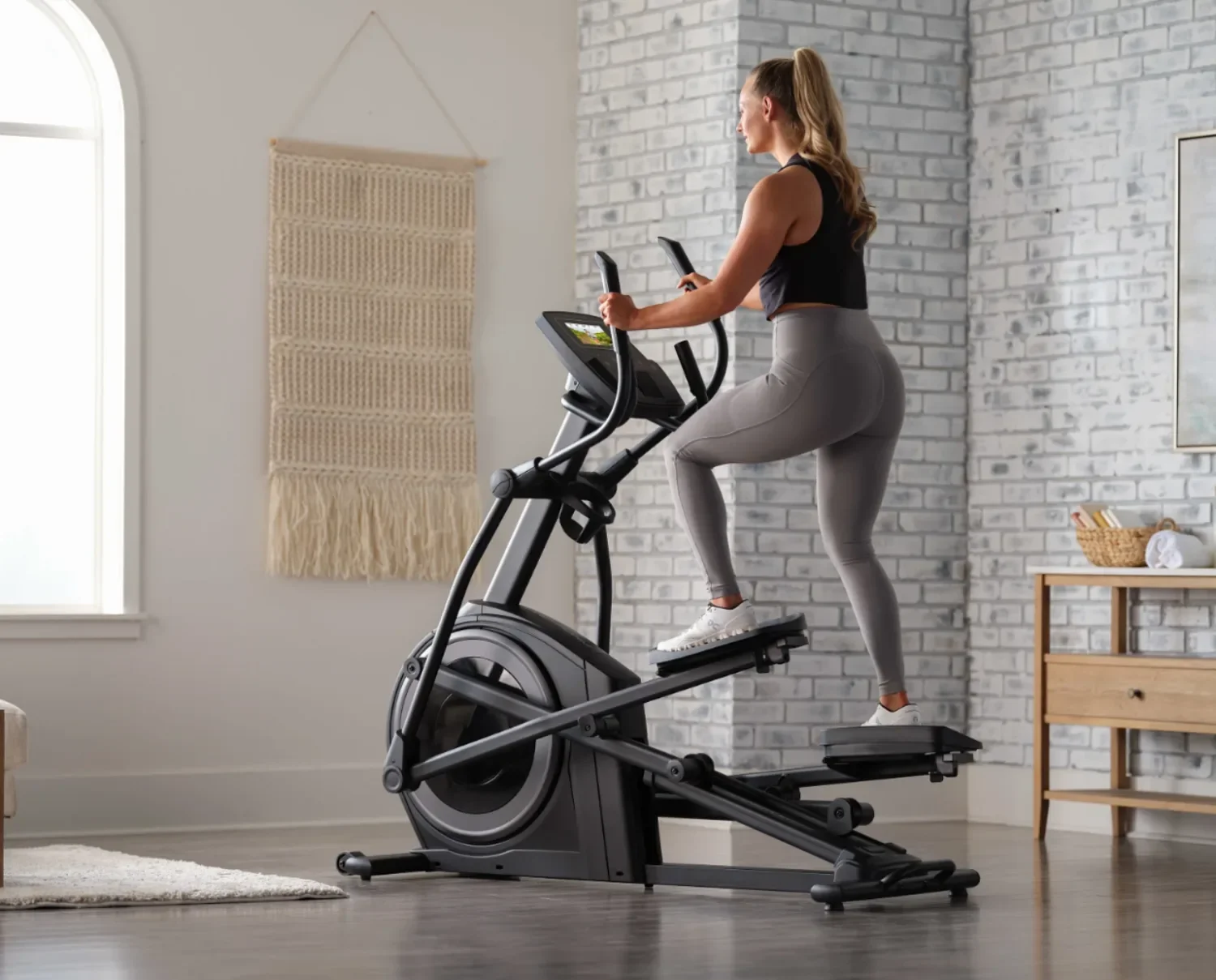 A woman in workout clothes exercising on an elliptical machine in a home gym with white brick walls and natural light.