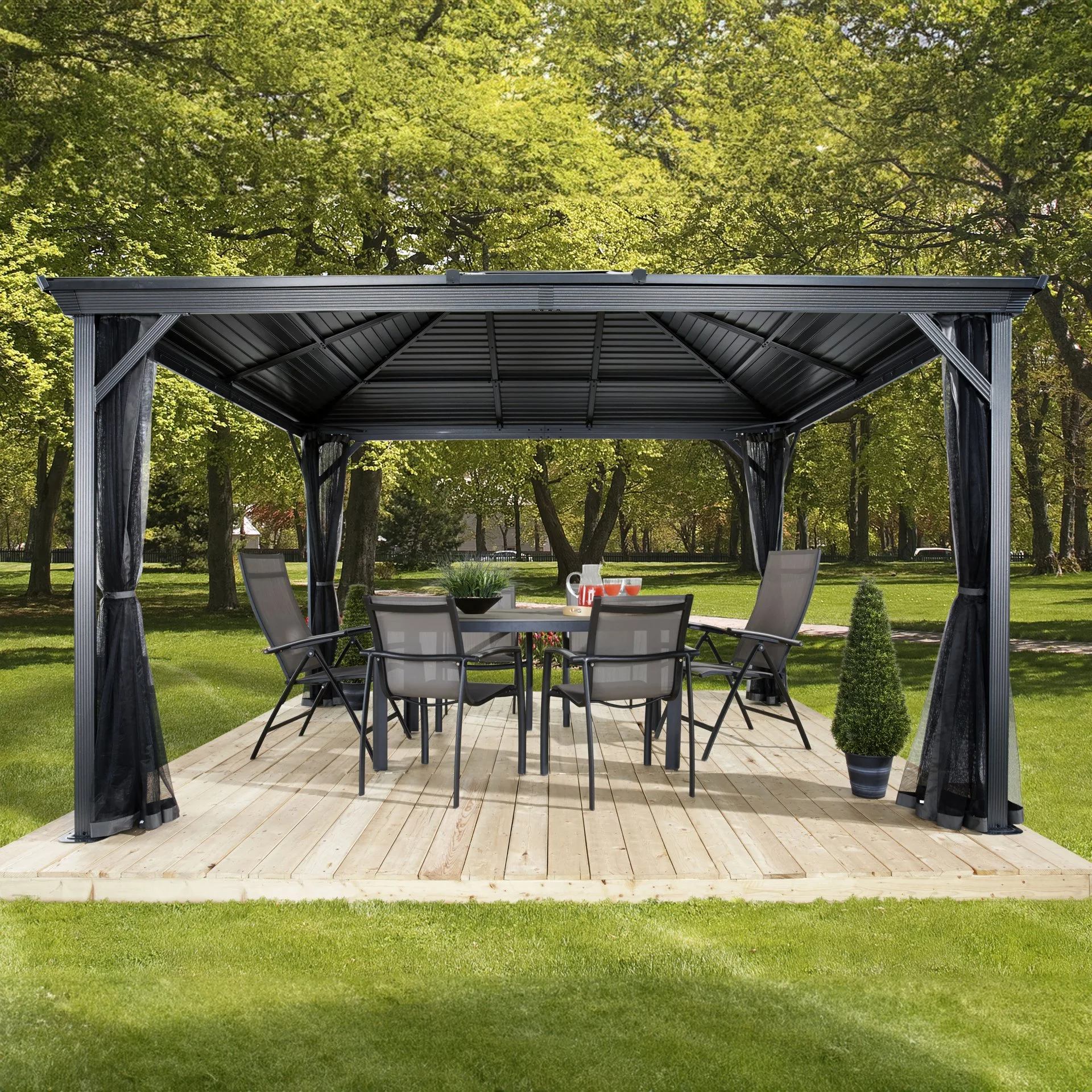 A black outdoor gazebo with curtains over a wooden deck, surrounding a table with six chairs, set in a lush green garden with trees and grass.
