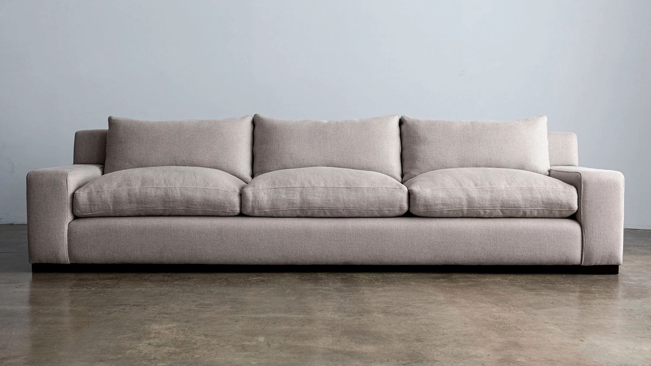 A light beige fabric sofa with three cushions, set against a plain light gray wall on a concrete floor.